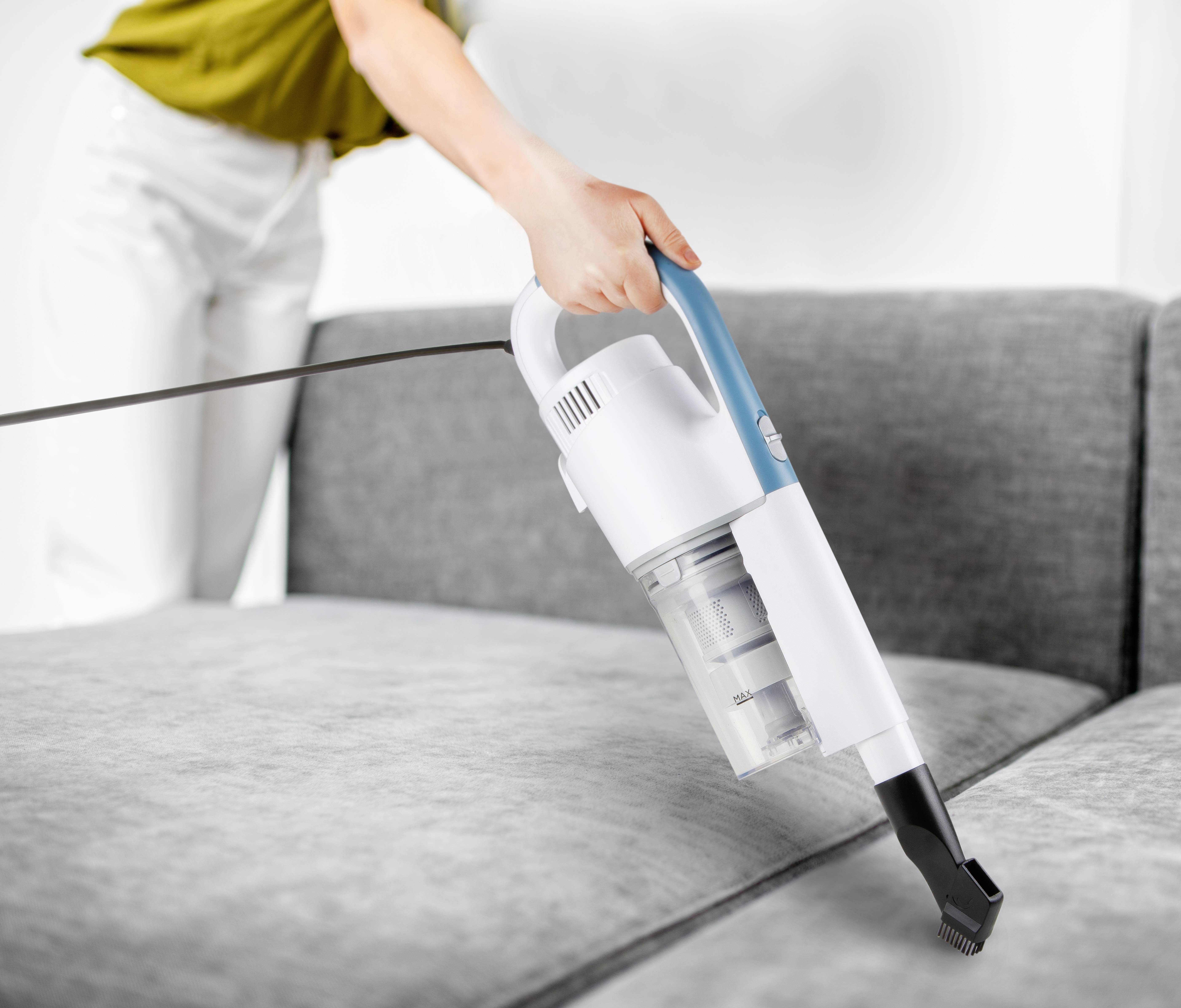 Eine Person reinigt ein graues Sofa mit einem weißen Handstaubsauger. Die Person trägt helle Kleidung.