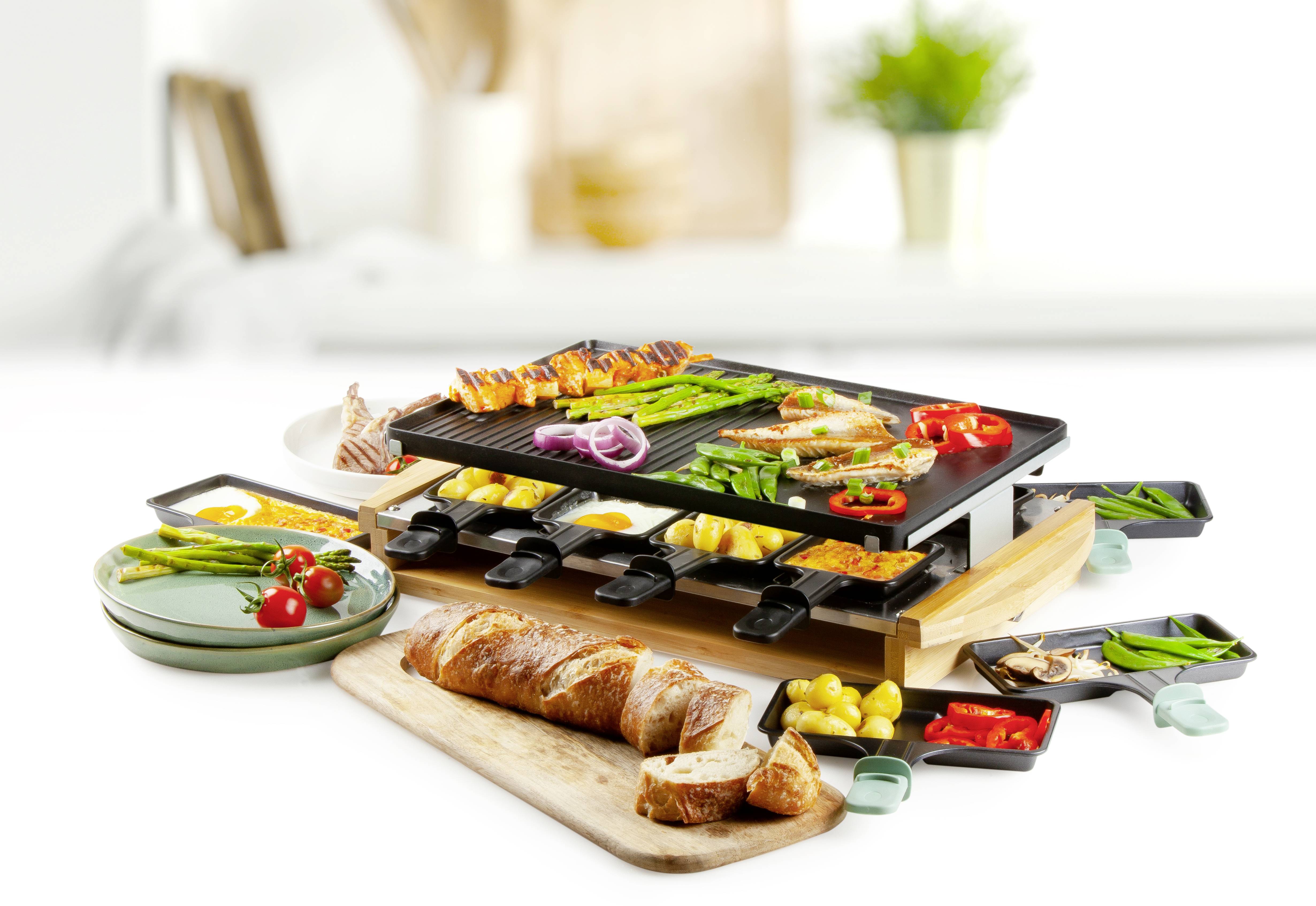 Ein Raclette-Grill mit verschiedenen Speisen wie Fleisch, Gemüse, Eiern und Käse. Im Hintergrund ist unscharf ein heller Raum zu sehen.