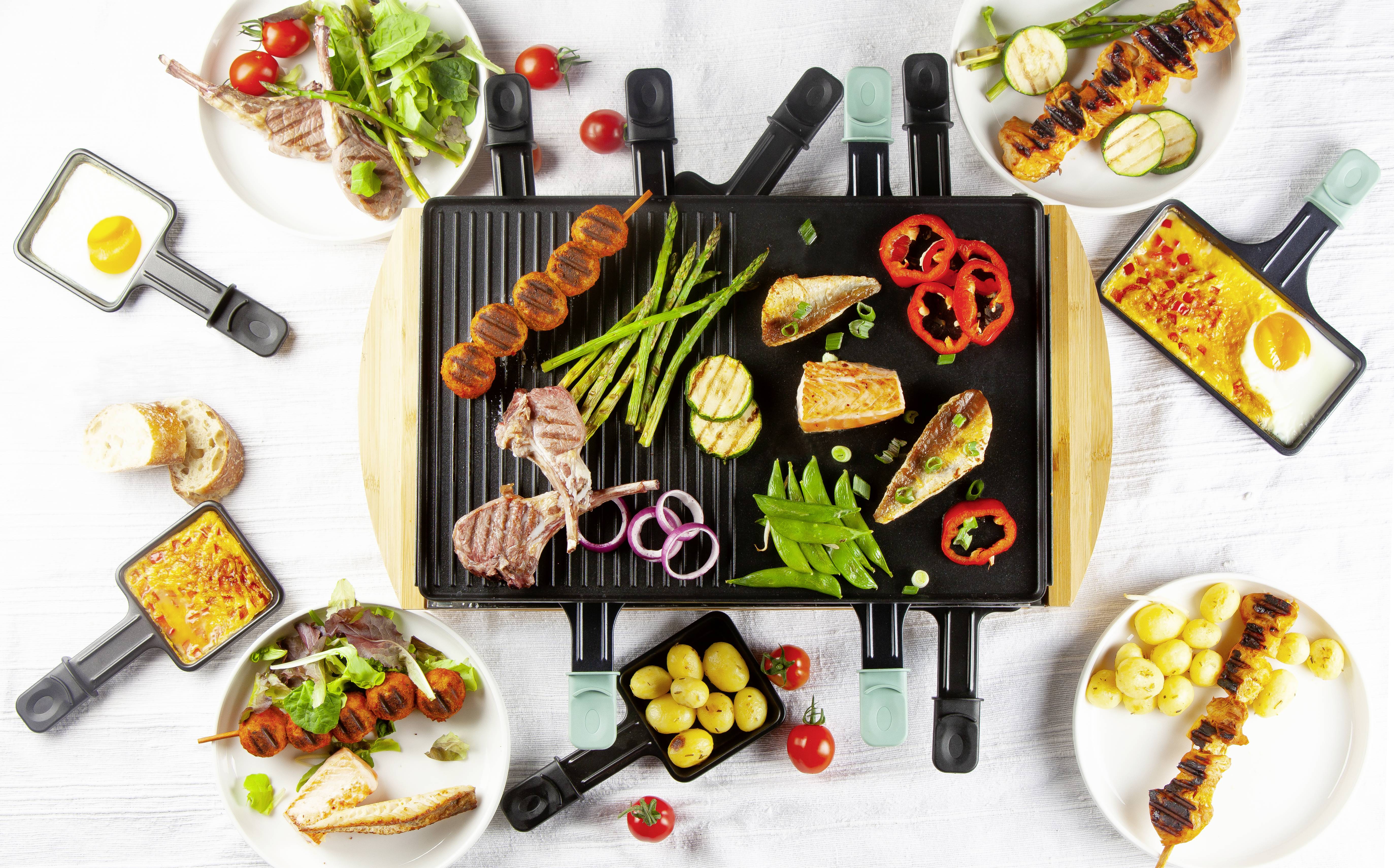 Ein Tisch mit Raclette-Grill in der Mitte, umgeben von kleinen Pfännchen mit Zutaten wie Kartoffeln, Gemüse und Fleisch.