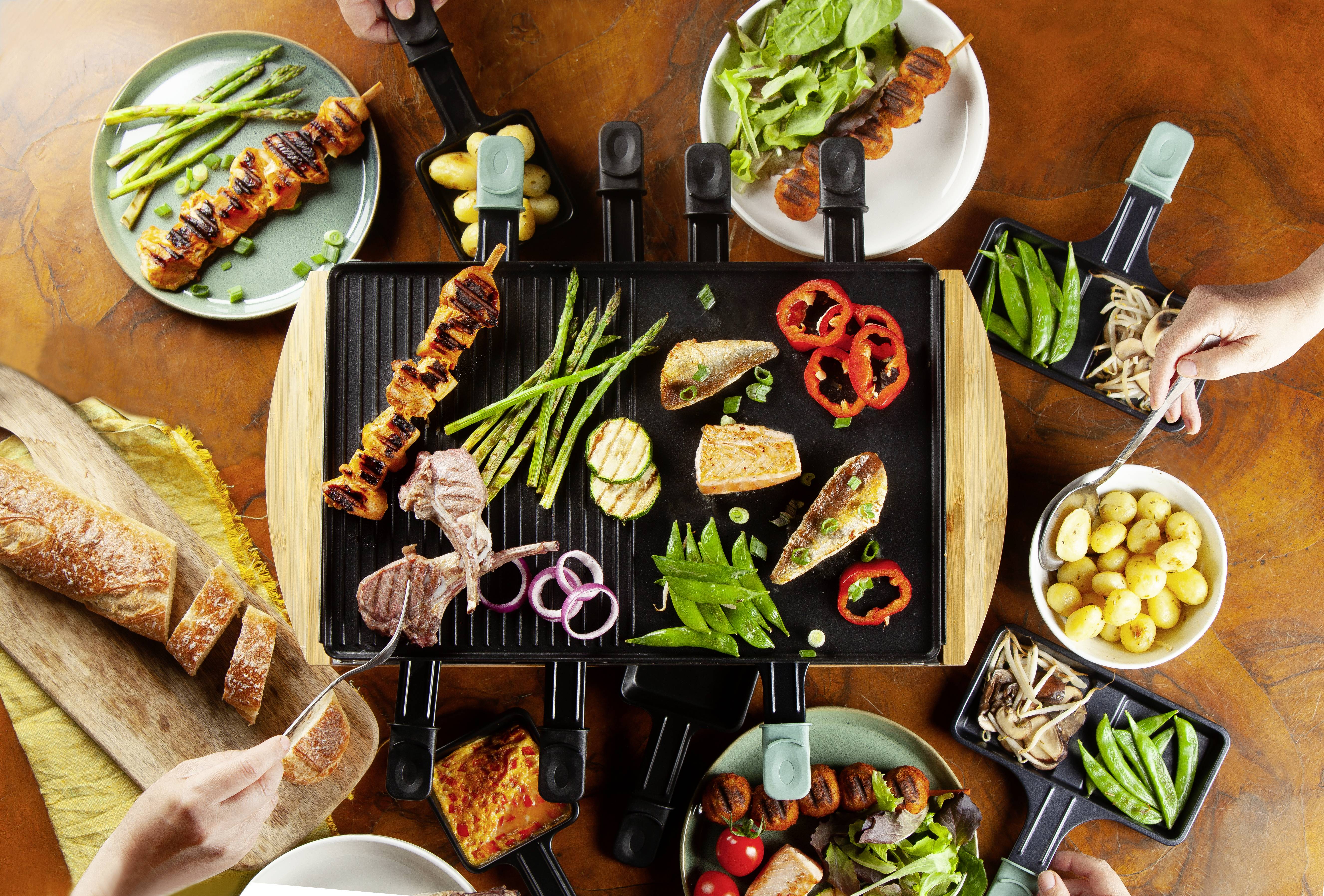 Ein Tisch mit gegrilltem Gemüse, Fleisch und Fisch auf einem Raclette-Grill, umgeben von Kräutern, Brot und Beilagen auf Tellern.