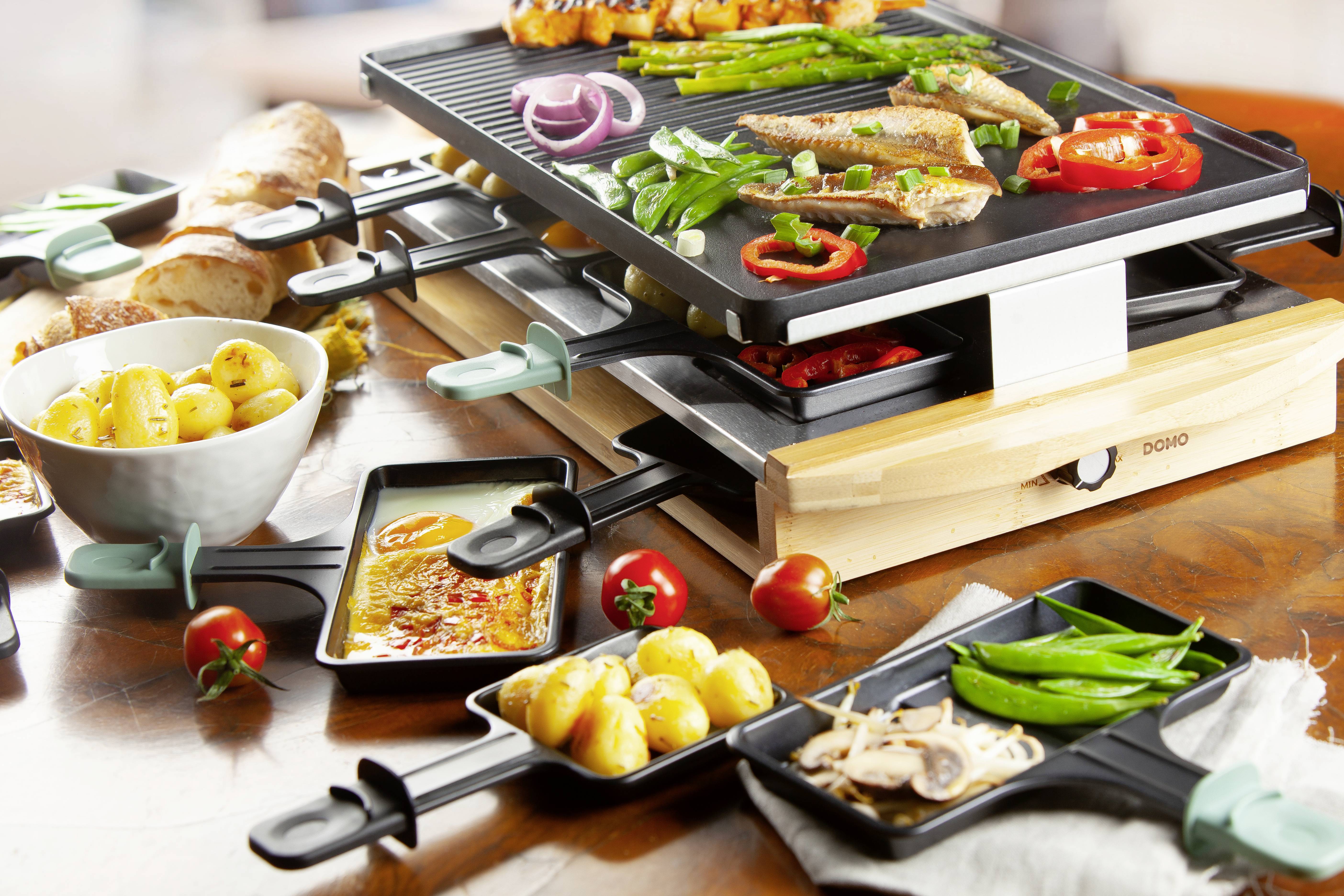Ein Raclette-Grill auf einem Tisch mit verschiedenen Grillpfännchen, die Gemüse, Fleisch und Käse enthalten. Im Hintergrund Brot und Tomaten.