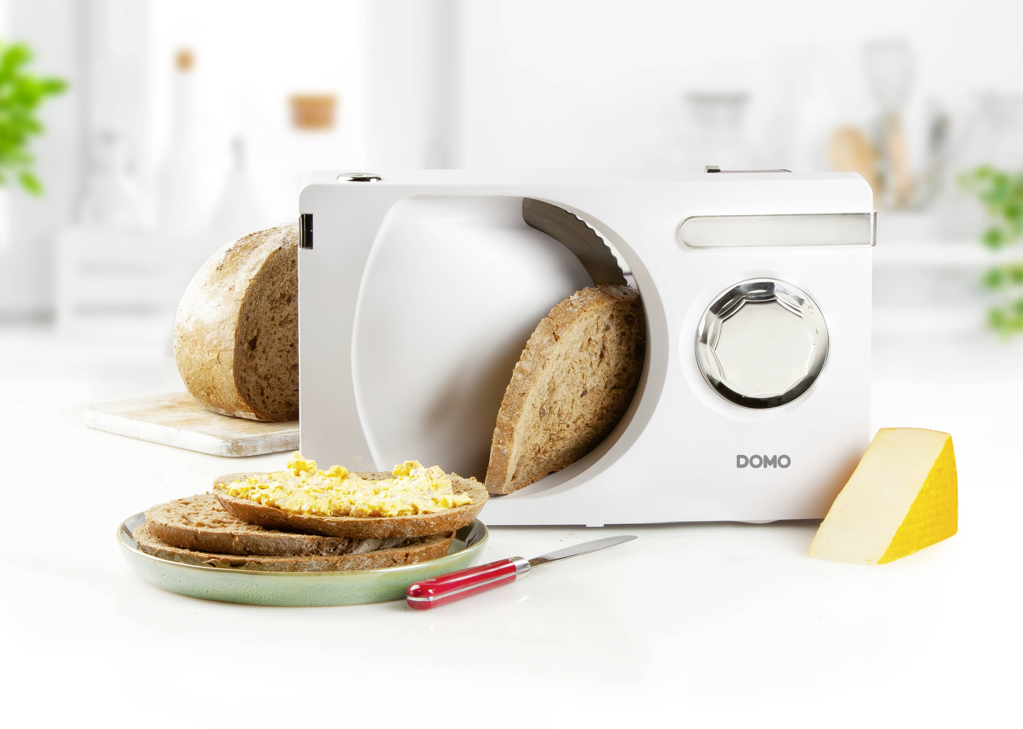 Ein elektrischer Allesschneider schneidet eine Scheibe Brot. Daneben liegt ein Teller mit bestrichenem Brot und einem Messer. Im Hintergrund unscharf.