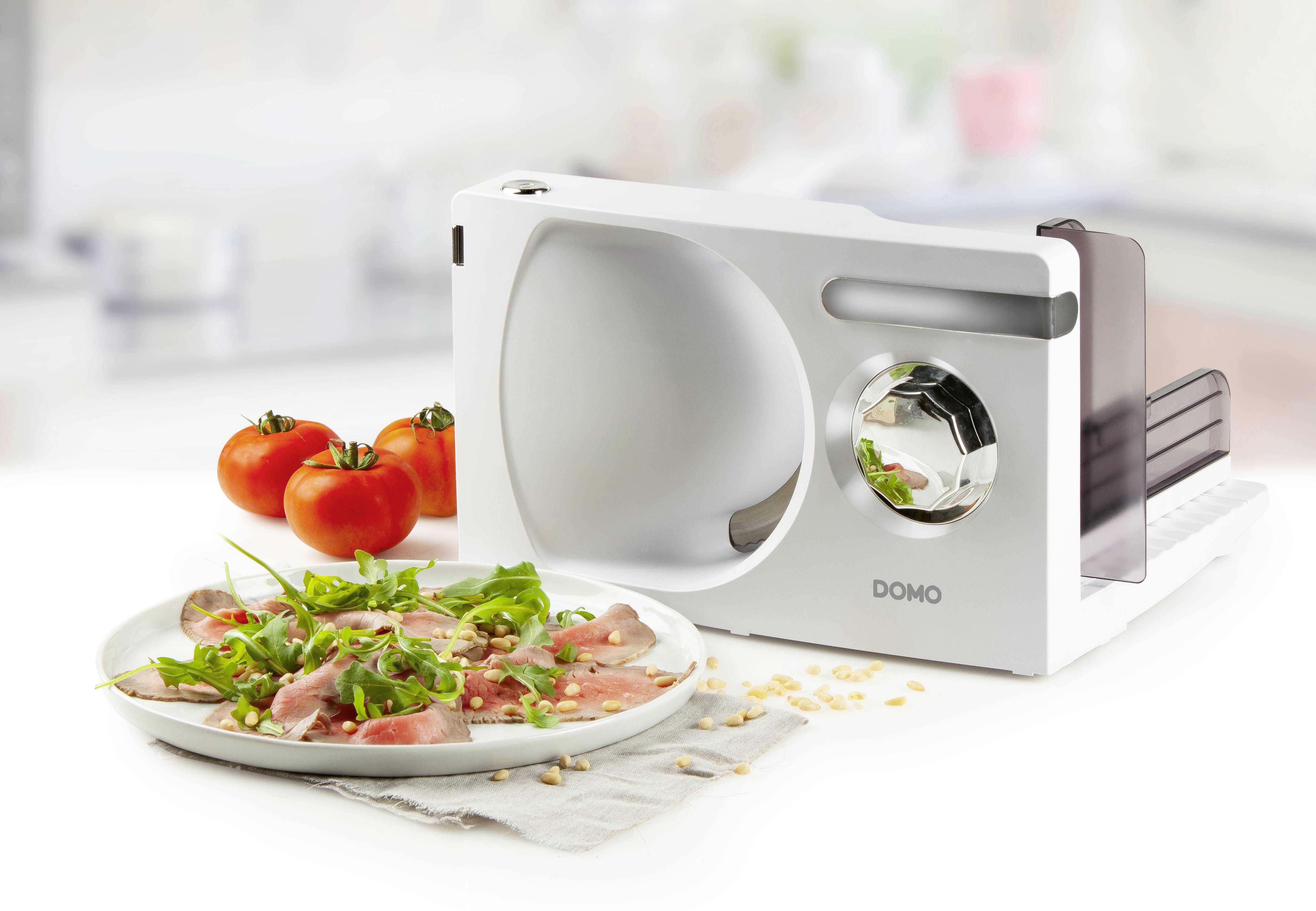 Elektrischer Allesschneider auf weißem Tisch, daneben Teller mit Carpaccio, Tomaten im Hintergrund. Perfekt für präzises Schneiden.