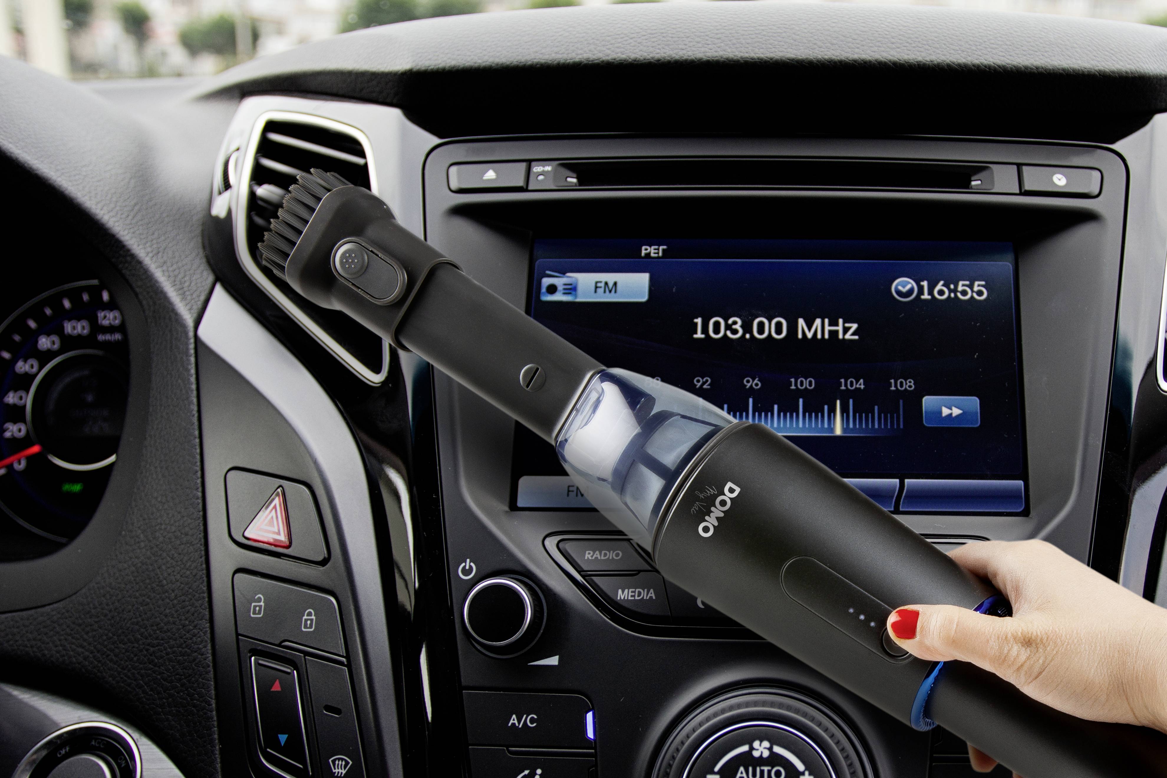 Eine Hand hält einen kleinen tragbaren Staubsauger, der das Armaturenbrett eines Autos reinigt. Im Hintergrund läuft das Radio.