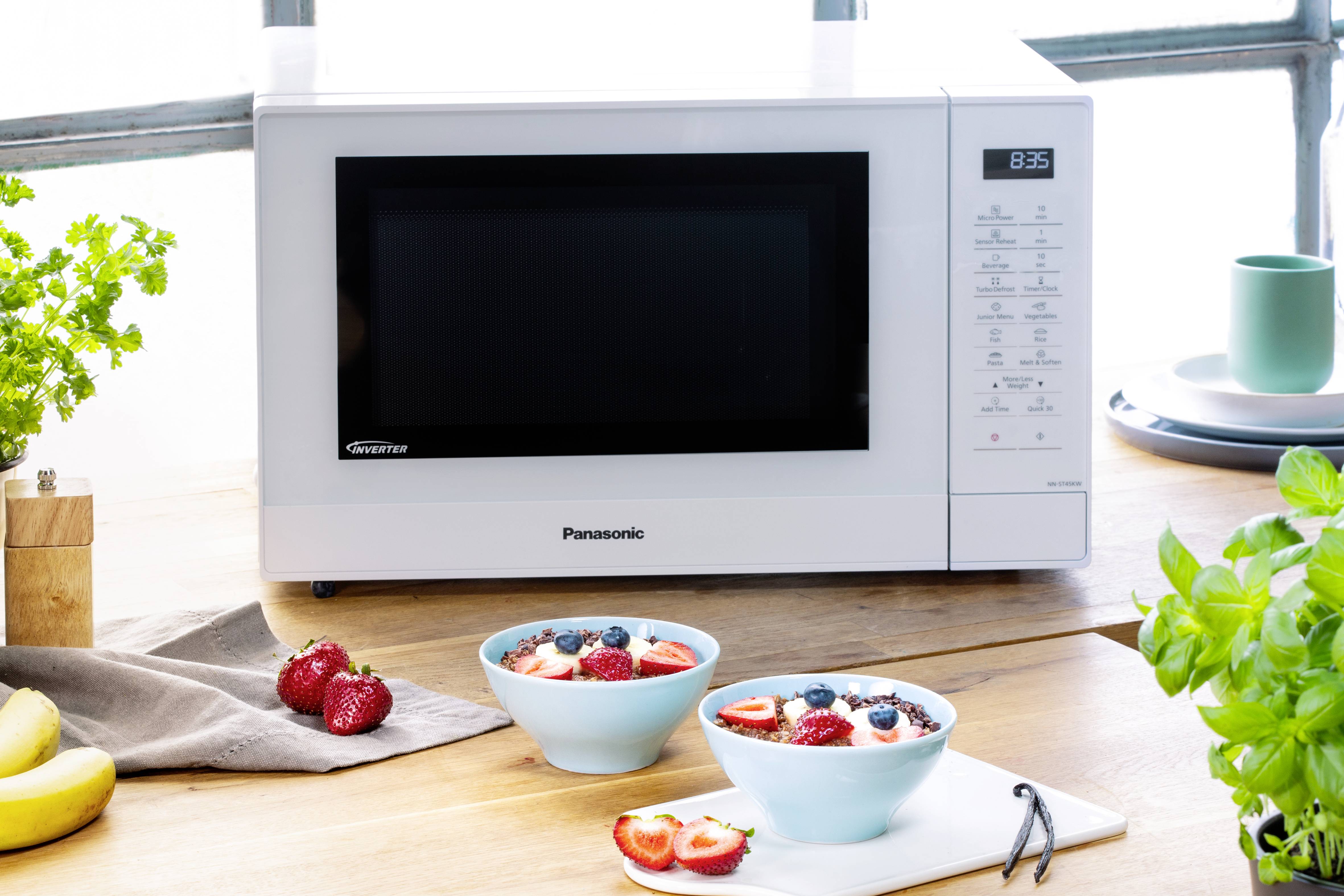 Eine weiße Mikrowelle steht auf einem hellen Holztisch, daneben zwei Schalen mit Joghurt und Beeren, umgeben von frischen Erdbeeren und Kräutern.