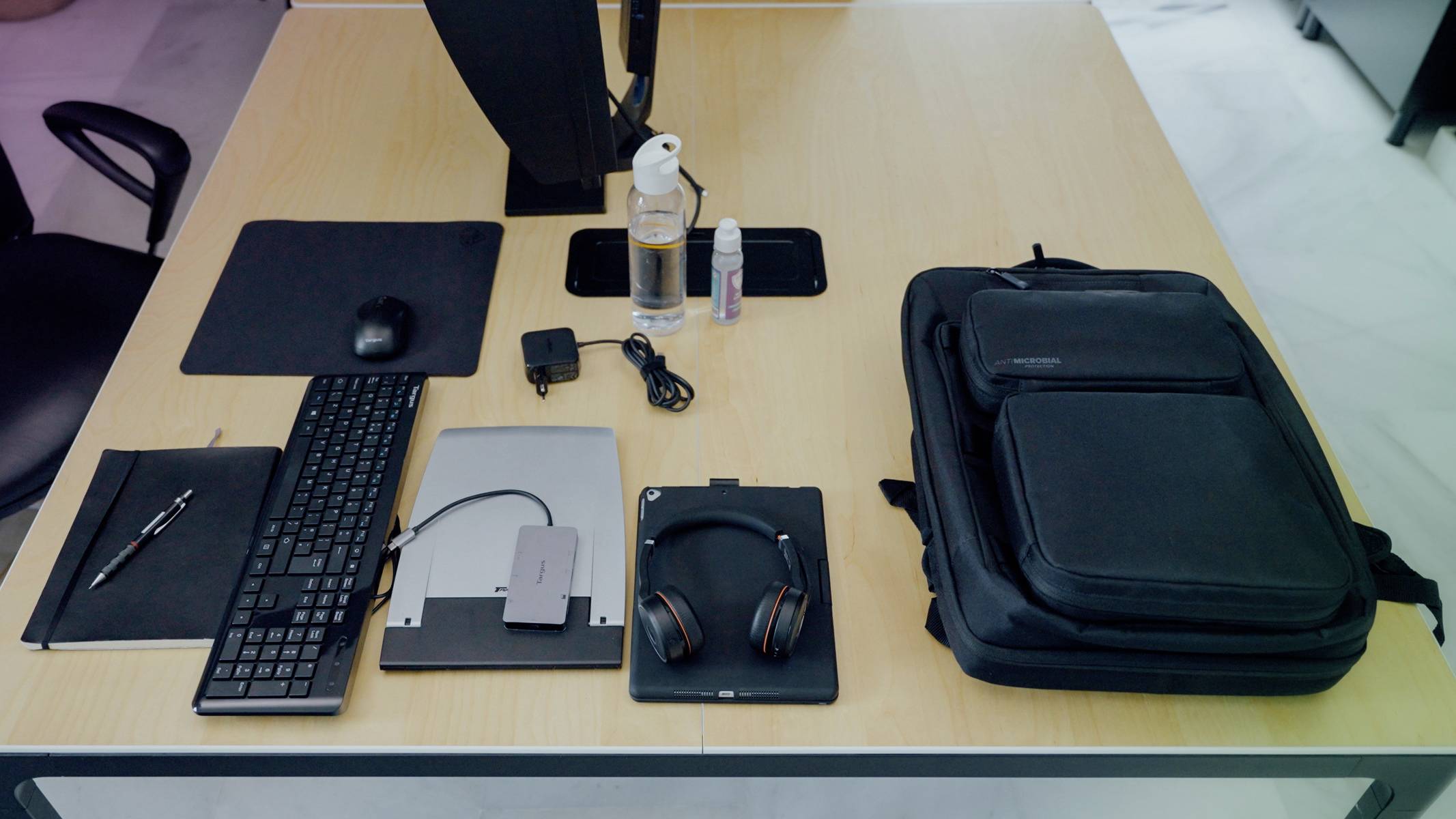 Ein aufgeräumter Schreibtisch mit Laptop, Tastatur, Mauspad, Kopfhörern, Stift, Rucksack, Wasserflasche und Desinfektionsmittel.