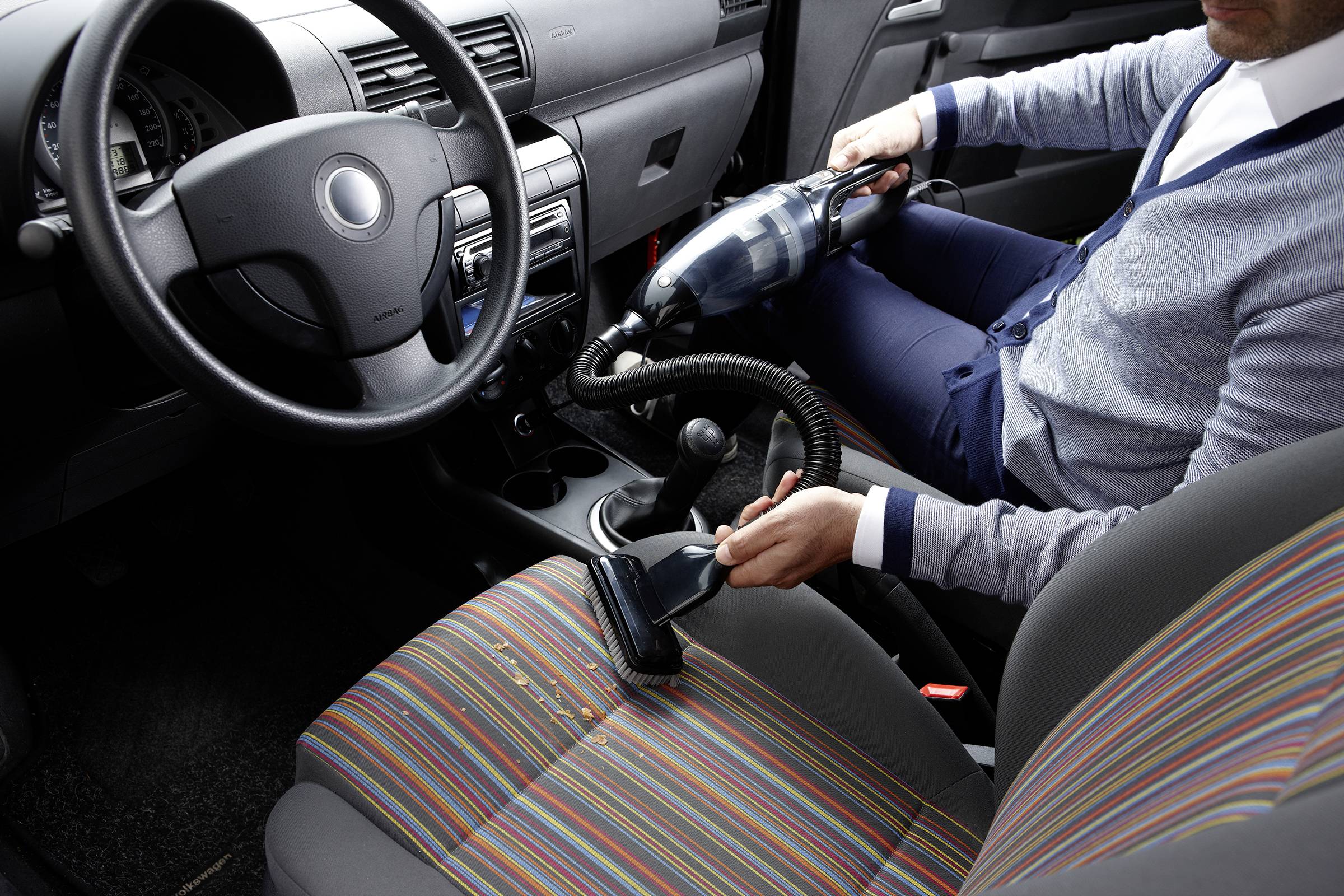 Eine Person saugt den Innenraum eines Autos mit einem Handstaubsauger. Der Fokus liegt auf der gründlichen Reinigung des Fahrzeugsitzes.