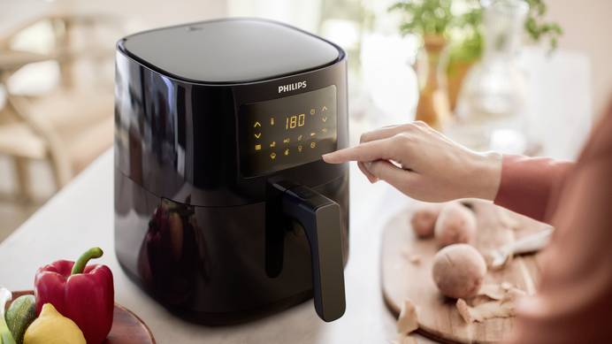 Eine Heißluftfritteuse von Philips steht auf einem Tisch. Neben ihr finden sich ein Teller mit einer Paprika, zwei Zucchinis und einer Zitrone sowie ein Schneidbrett, auf dem Kartoffeln liegen. Abgeschälte Schale und ein Kartoffelschäler weisen darauf hin, dass jemand begonnen hat, die Kartoffeln zu schälen. Eine Person, von der man nur die rechte Hand, den rechten Unterarm und unscharf den linken Oberarm sieht, bedient die Heißluftfritteuse. Auf dem Display sind die Zahl 180 (steht für die eingestellte Temperatur) und diverse Icons für unterschiedliche Optionen zu sehen.