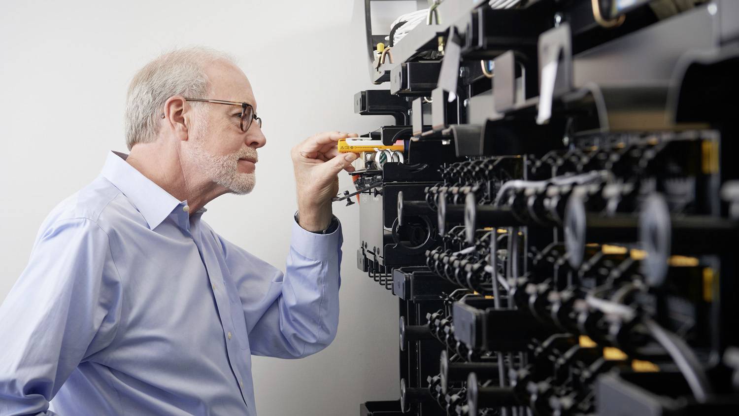 Ein älterer Mann inspiziert aufmerksam ein großes Regal mit digitalen Kabeln und Geräten, möglicherweise in einem technischen Umfeld.