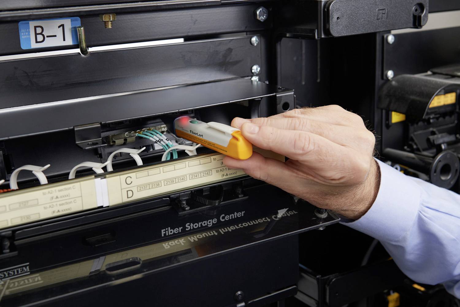 Eine Person testet mit einem optischen Stift ein Glasfaser-Verbindungspanel in einem Serverraum. Links sind Etiketten mit Buchstaben.