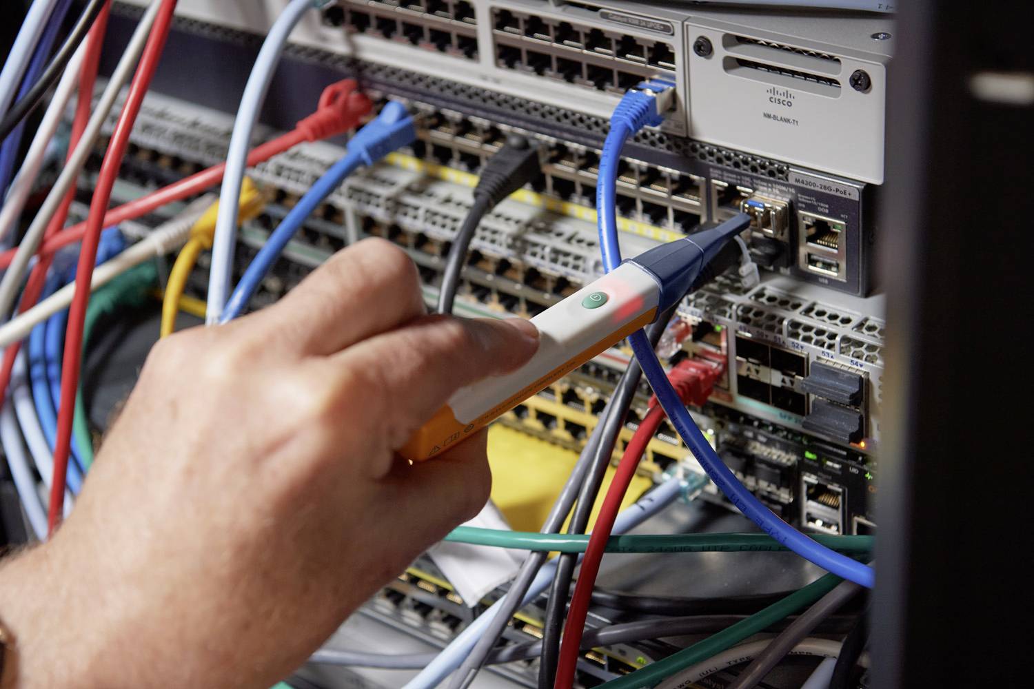 Eine Hand hält ein Prüfwerkzeug und berührt Netzwerkkabel an einem voll belegten Server-Rack, um Netzwerkkonnektivität zu testen.