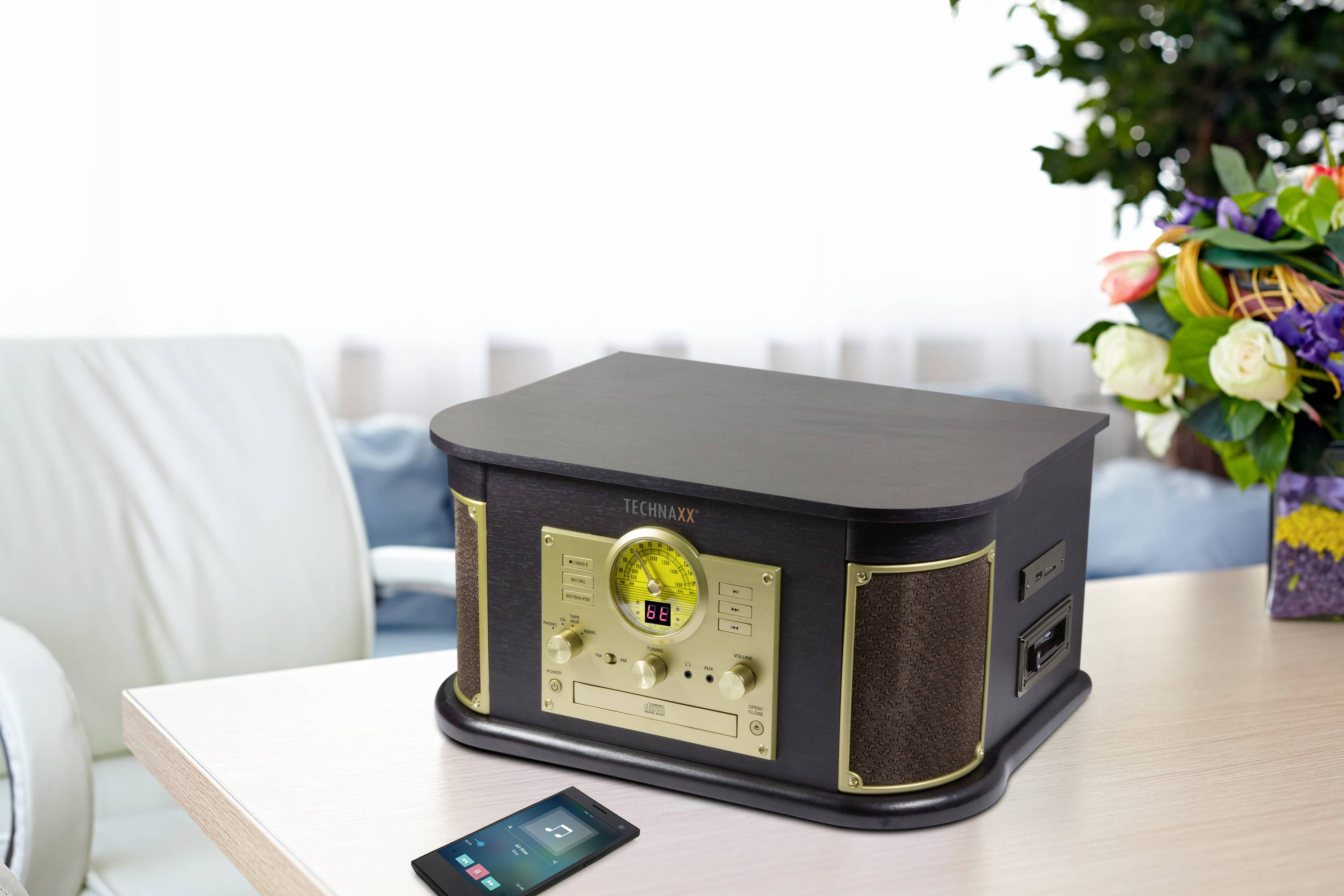 Ein Retro-Stereo-Radio mit Holzgehäuse auf einem Tisch. Daneben liegt ein Smartphone. Im Hintergrund sind Blumen zu sehen.
