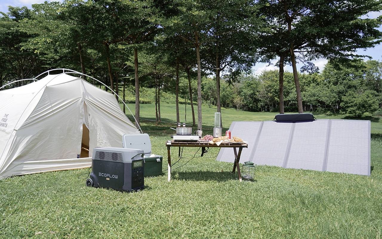 Zelt und Campingausrüstung auf grüner Wiese, umgeben von Bäumen. Tisch mit Lebensmitteln, Solarpanel und Kühlbox im Vordergrund.