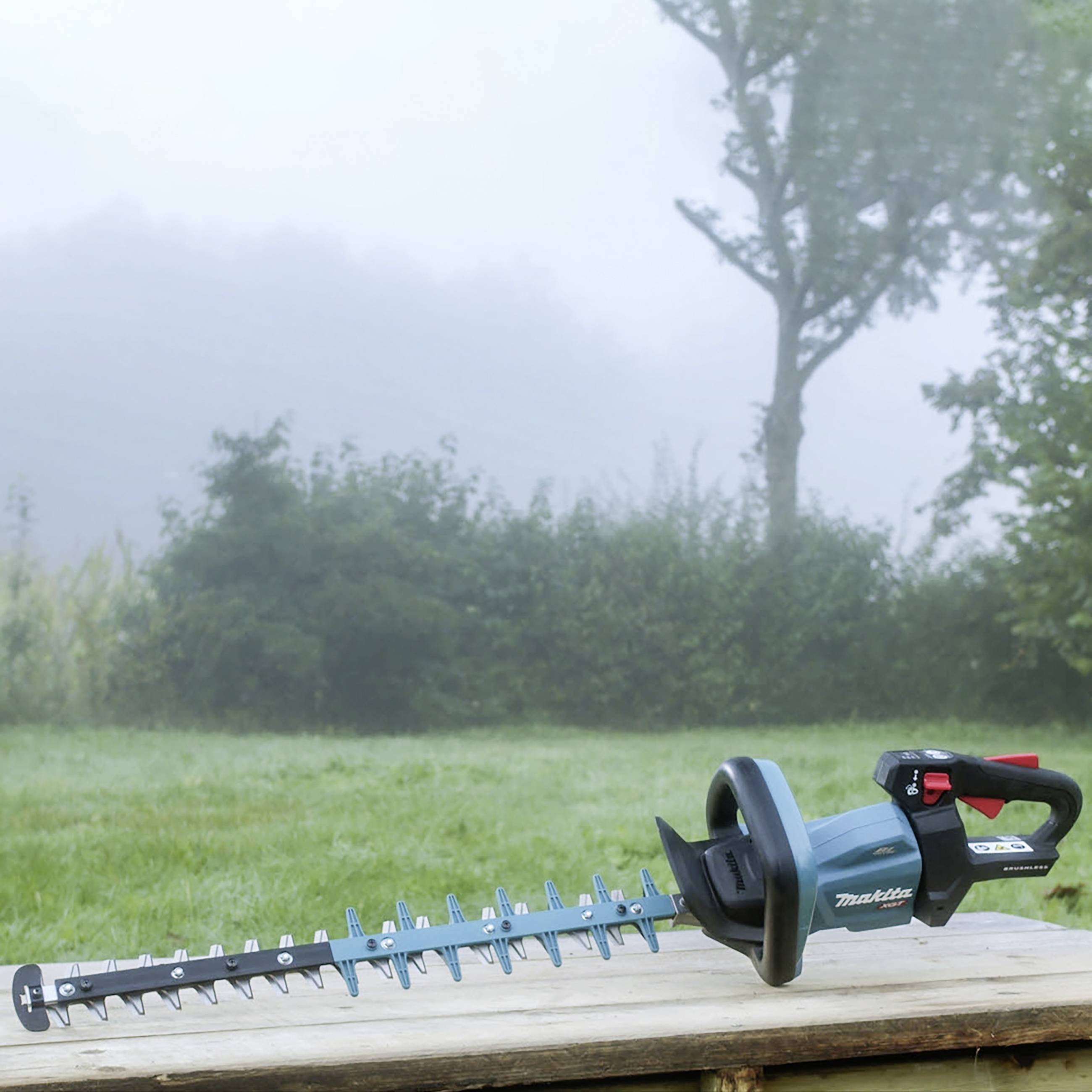 Heckenschere liegt auf einem Holztisch im Freien, mit nebliger Landschaft und Bäumen im Hintergrund.