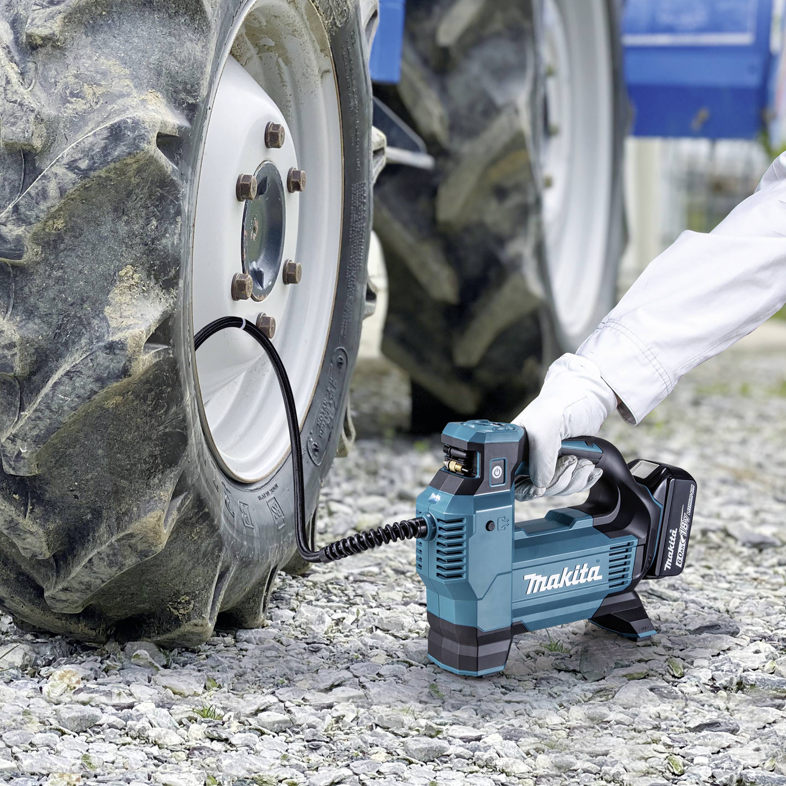 Eine Person pumpt einen Traktorreifen mit einem blauen, tragbaren Kompressor auf. Der Reifen steht auf einem Kiesuntergrund.