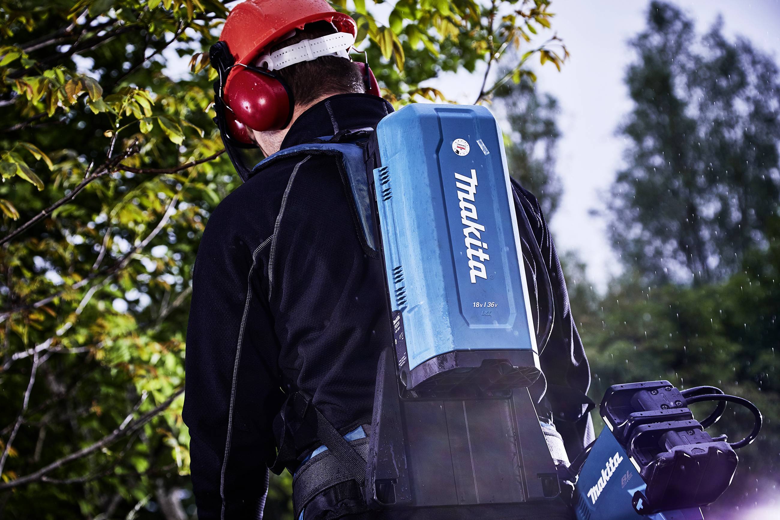Ein Mann trägt einen blauen Makita-Akkupack und Ohrenschützer im Freien, umgeben von Bäumen.