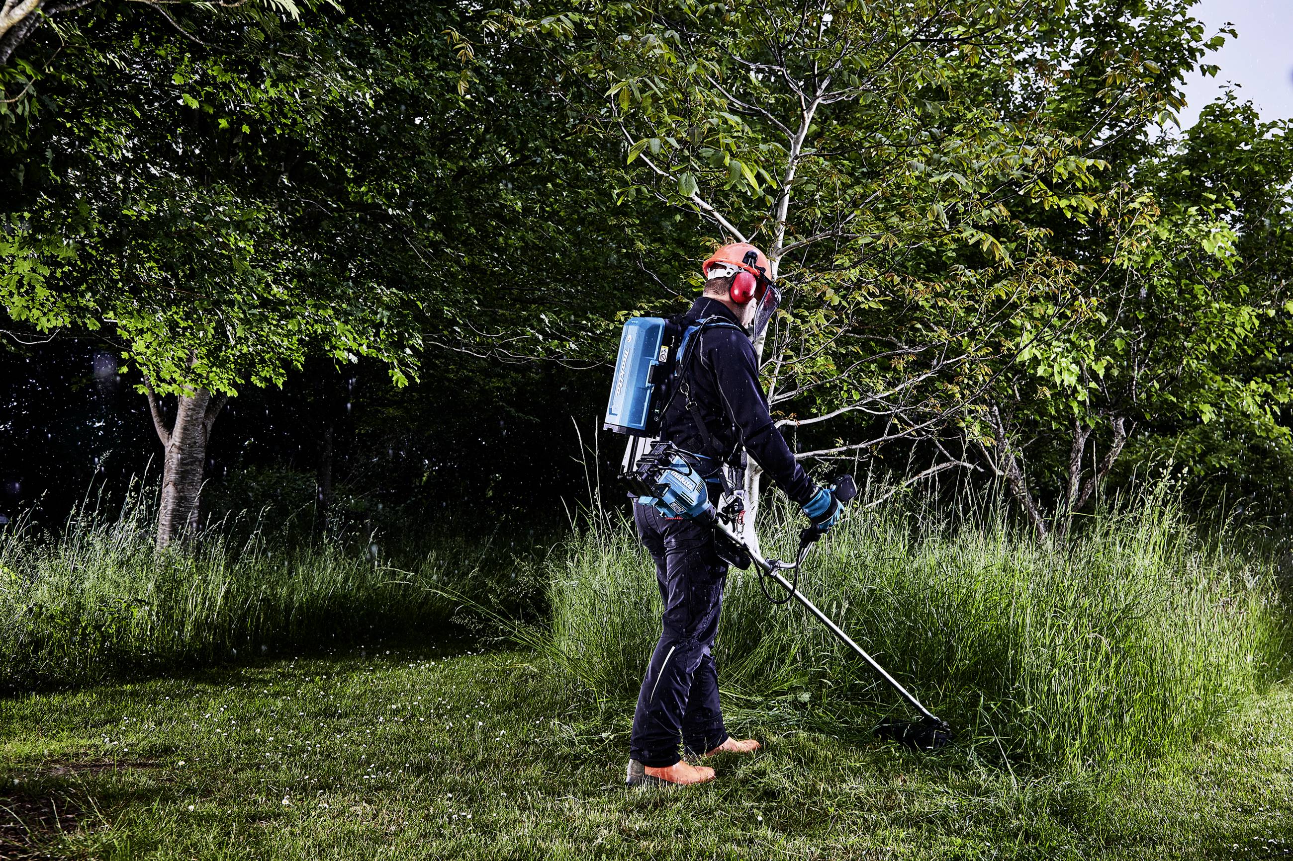 Eine Person in Schutzkleidung trimmt Gras mit einem Motorsensen im Garten. Bäume im Hintergrund bieten eine grüne Kulisse.
