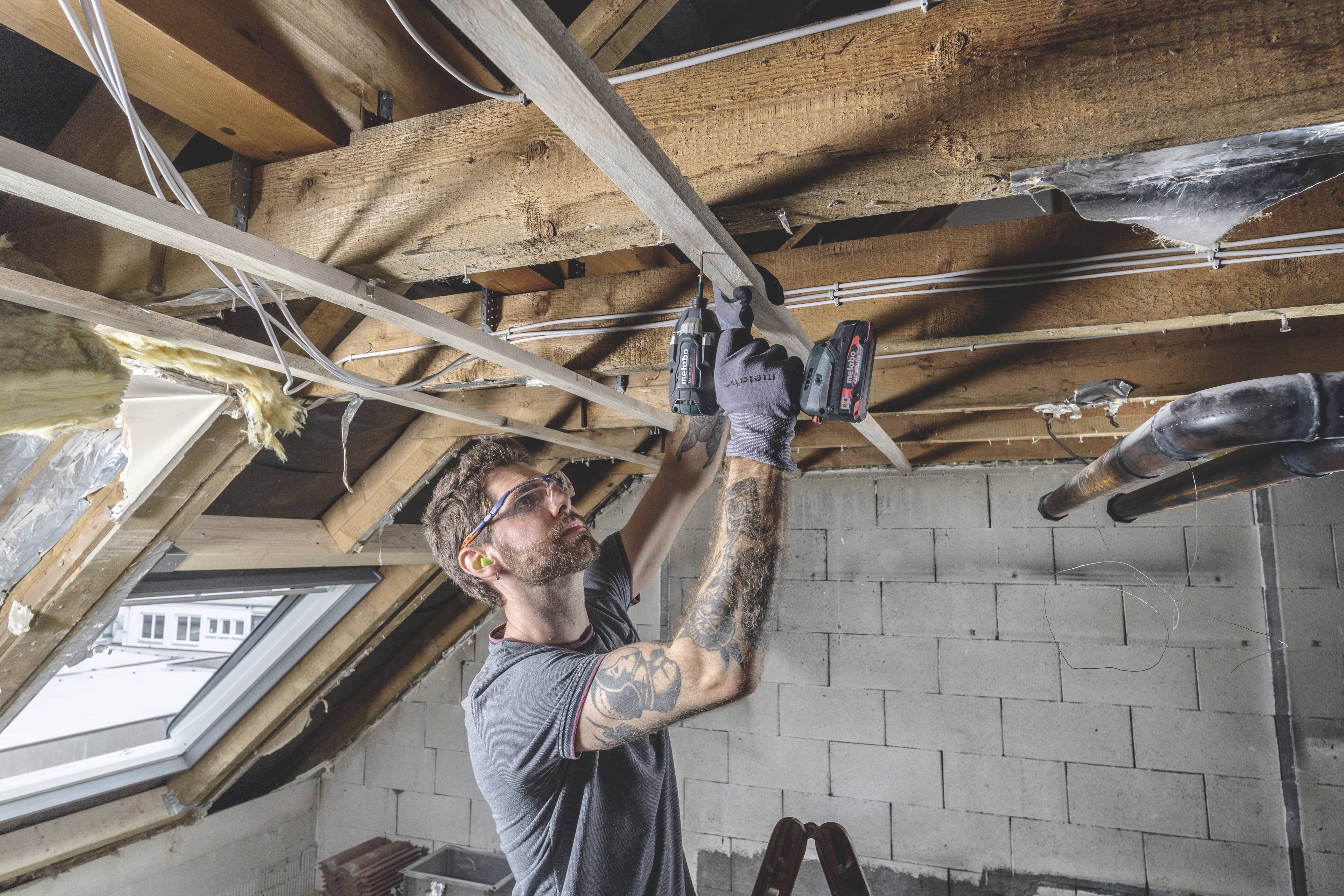 Ein Handwerker arbeitet mit einem Akkuschrauber an Holzbalken in einem Dachboden. Er trägt Schutzbrille und Arbeitshandschuhe.