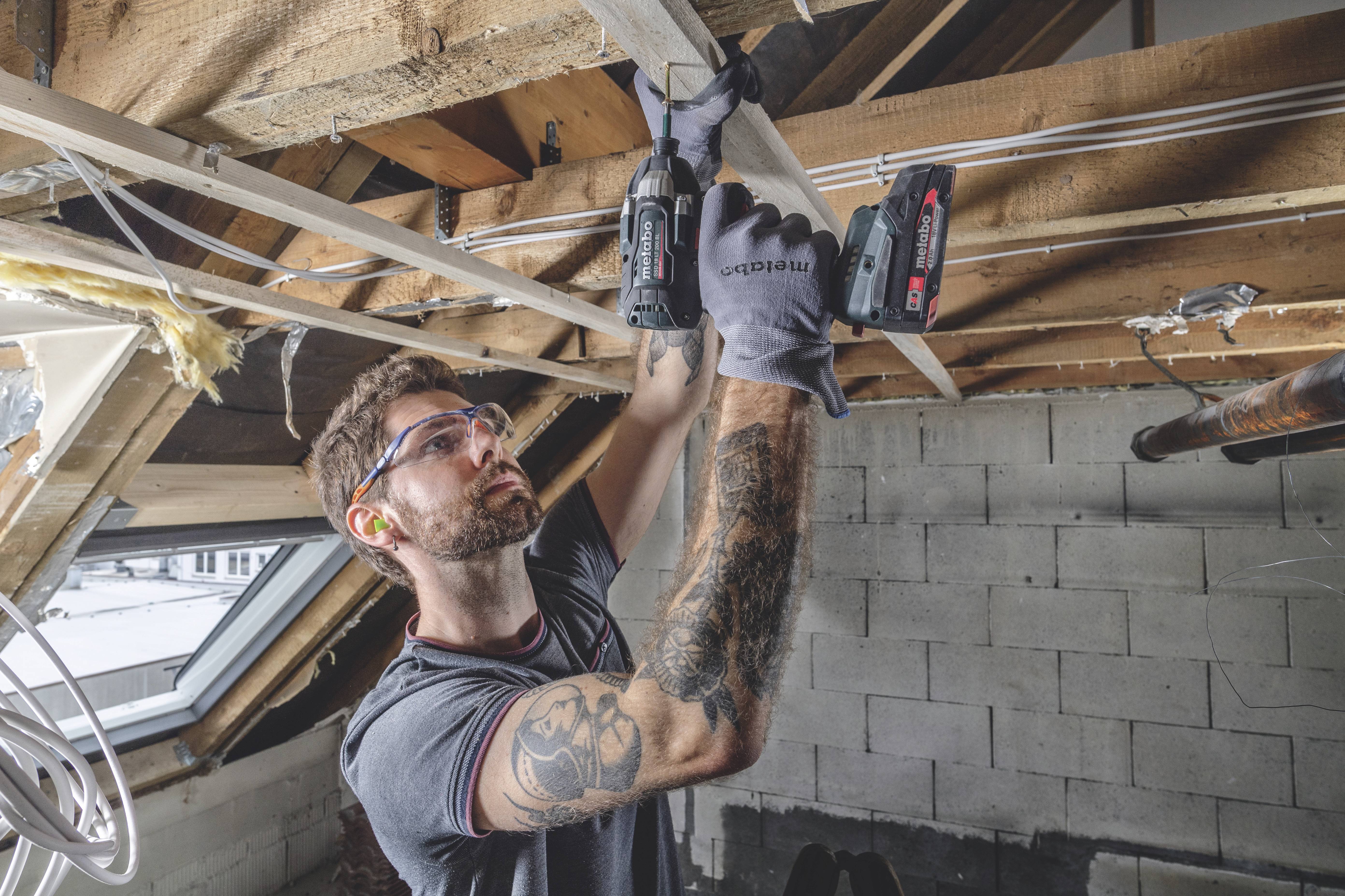 Ein Mann mit Kurzarm-T-Shirt und Tattoos arbeitet mit einer Bohrmaschine an Holzbalken in einem Dachboden. Er trägt Sicherheitsbrille und Gehörschutz.