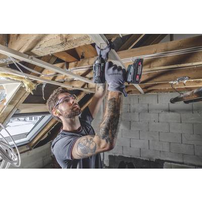 Ein Mann mit Kurzarm-T-Shirt und Tattoos arbeitet mit einer Bohrmaschine an Holzbalken in einem Dachboden. Er trägt Sicherheitsbrille und Gehörschutz.