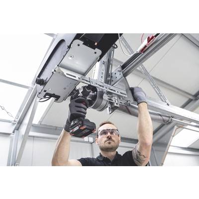 Ein Mann repariert eine Garagentür. Er trägt eine Schutzbrille und verwendet ein elektrisches Werkzeug, in einer Werkstattumgebung.