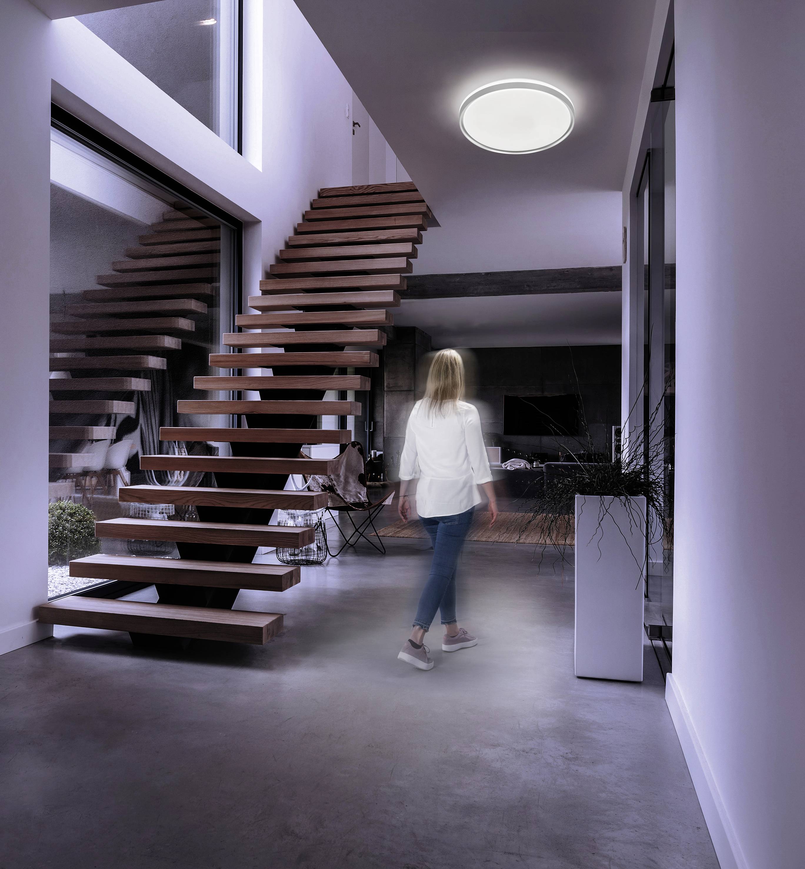 Eine Frau geht in einem modernen, minimalistischen Haus mit Holztreppe. Oberlichter und ein rundes Deckenlicht erhellen den Raum.