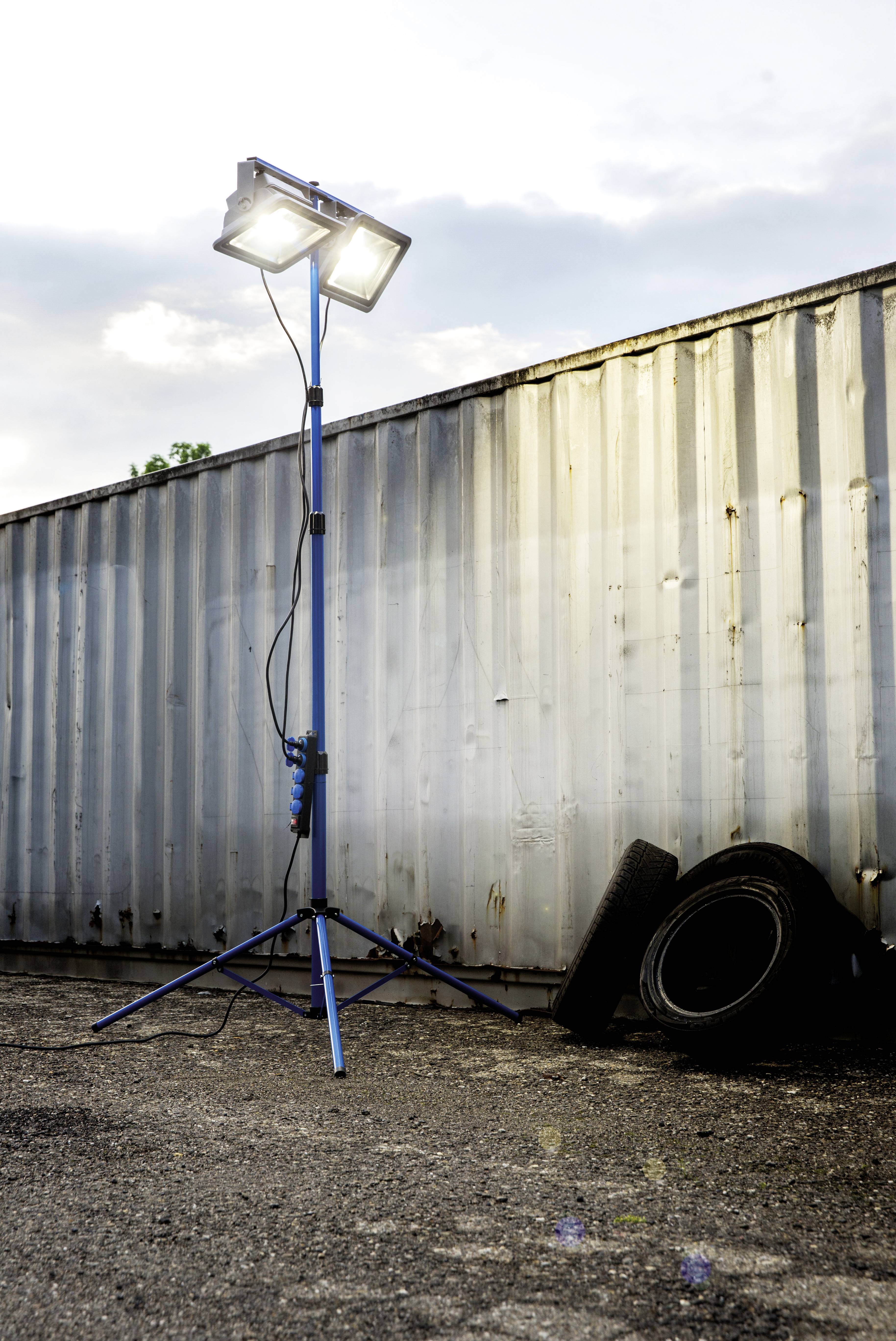 'Ein heller Baustrahler steht auf einem Dreibeinstativ neben einem Container. Daneben liegen zwei Autoreifen auf dem Boden.'