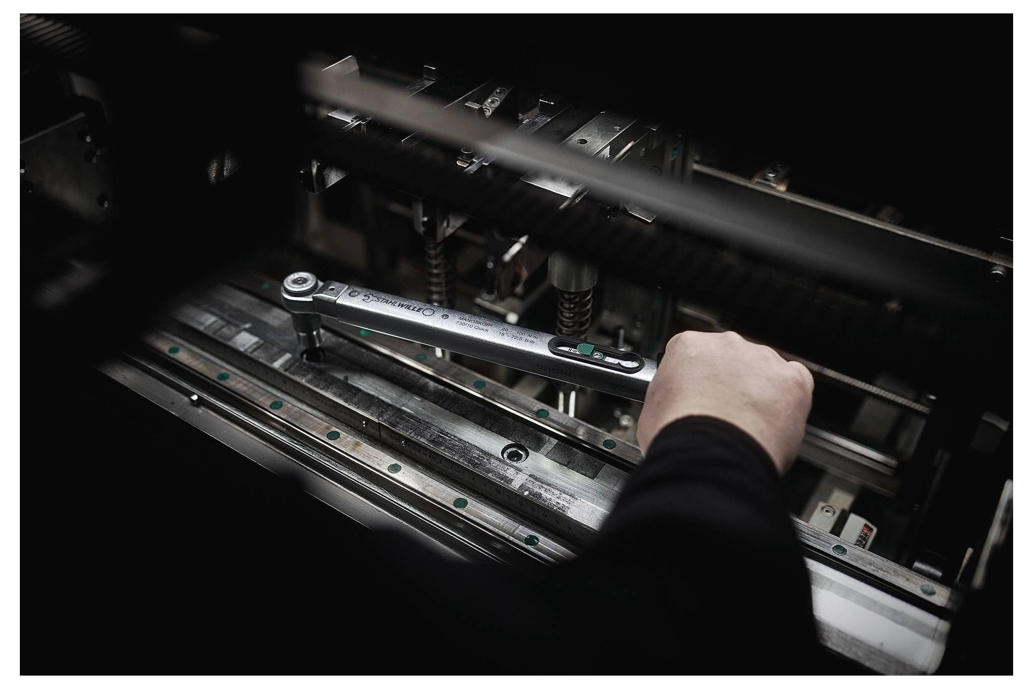 Eine Mechanikerin oder ein Mechaniker verwendet einen Schraubenschlüssel, um eine Maschine einzustellen. Der Fokus liegt auf Präzision und technischem Geschick in einer schwach beleuchteten Werkstatt.