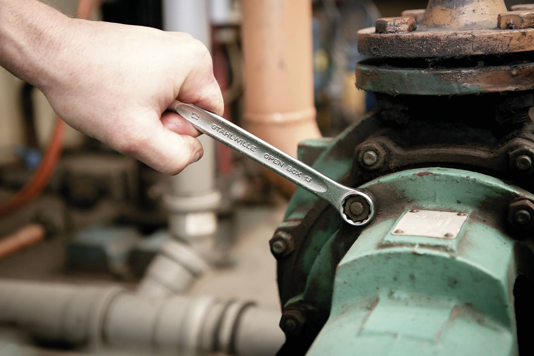 Eine Hand zieht mit einem Schraubenschlüssel eine Schraube an einer grünen Industriemaschine fest, was Wartungs- oder Reparaturarbeiten in einem mechanischen Umfeld andeutet.