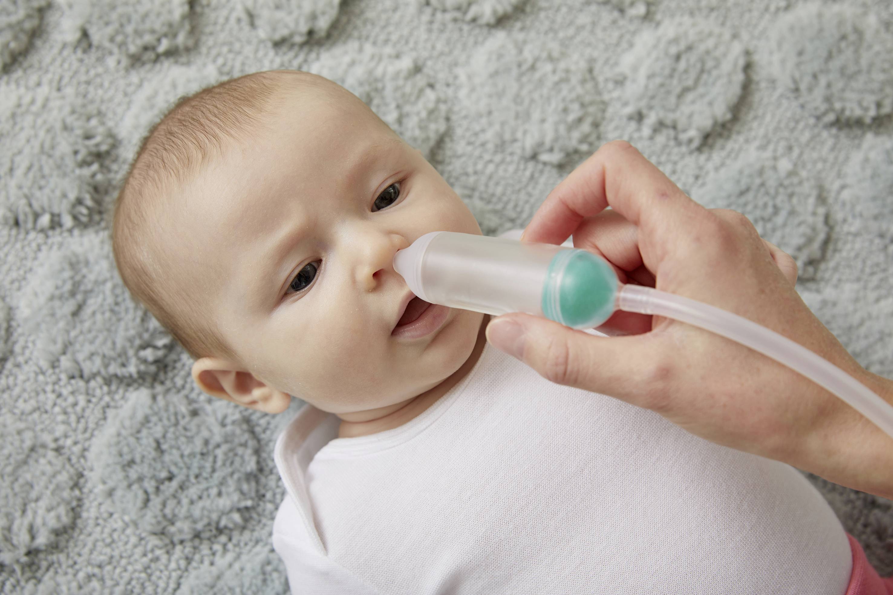 Ein Baby liegt auf einer Decke, während eine Hand einen Nasensauger sanft zur Reinigung der Nase des Babys verwendet.