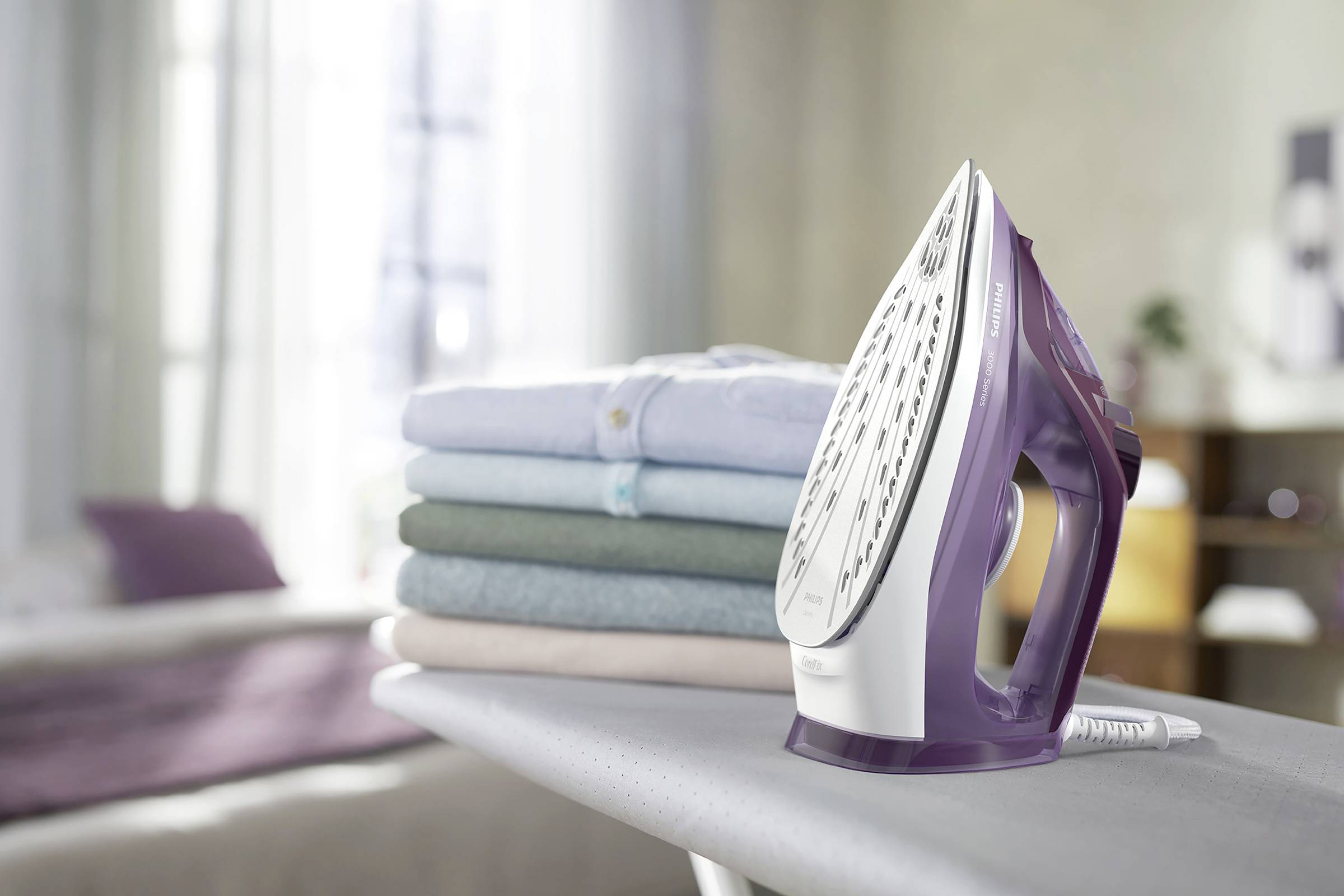 Bügeleisen auf einem Bügelbrett neben einem Stapel bunter, gefalteter Wäsche im Hintergrund. Sonnenlicht fällt auf die Szene.