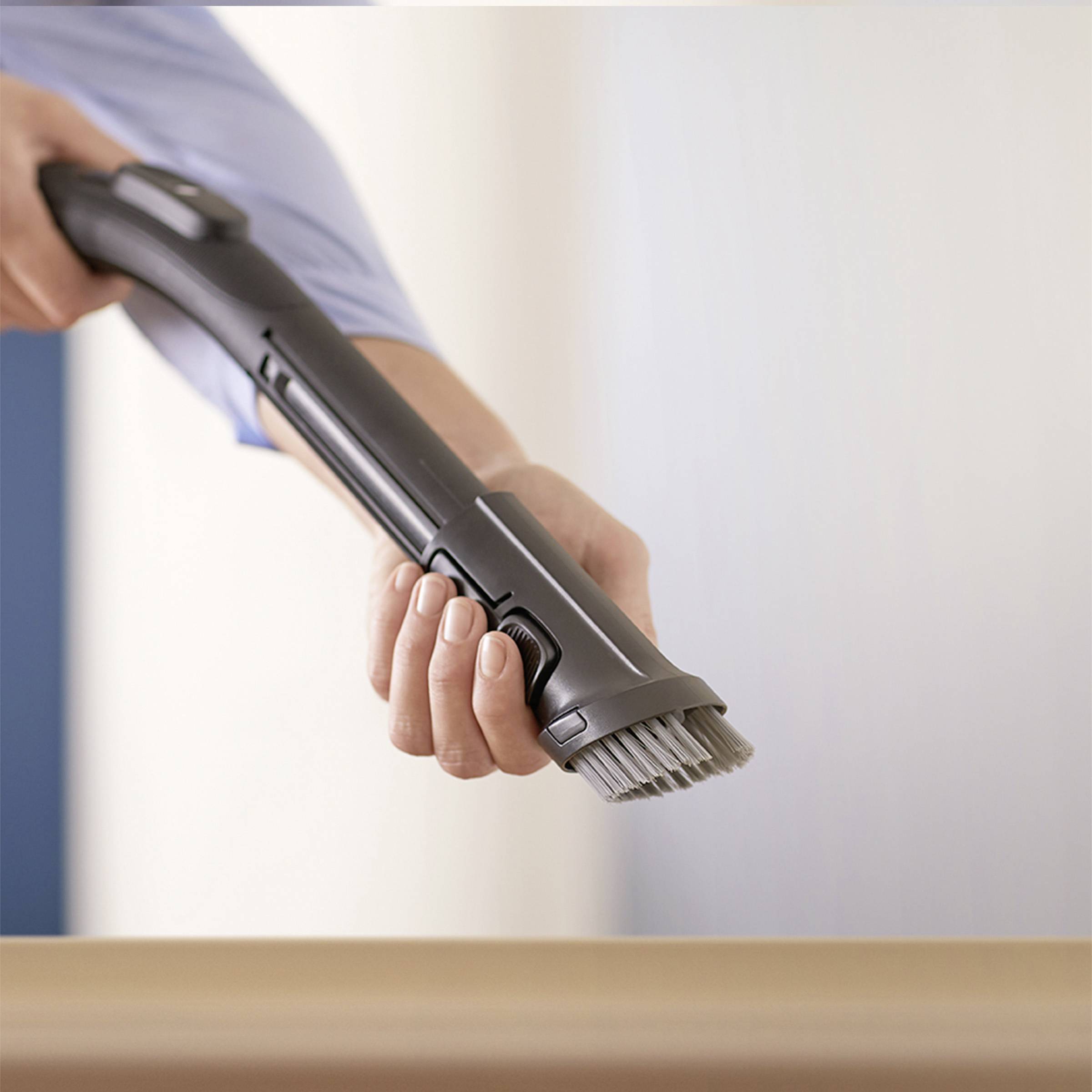 Eine Hand hält einen Staubsaugeraufsatz mit Bürste. Der Staubsauger wird über eine helle Oberfläche geführt.