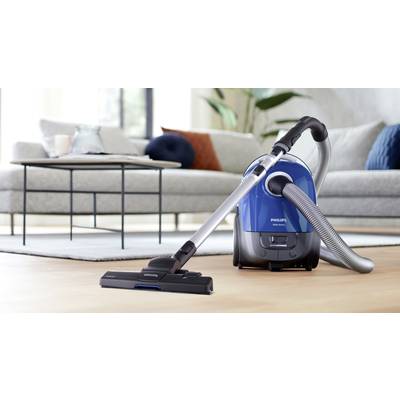 Ein blauer Staubsauger steht eingeschaltet auf einem Holzboden in einem Wohnzimmer mit grauem Sofa und Couchtisch im Hintergrund.