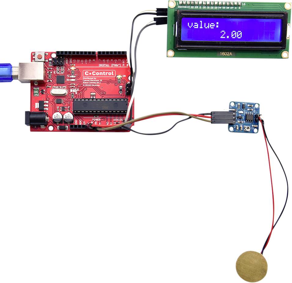 Eine rote Mikrocontroller-Platine ist mit einem blauen USB-Port verbunden. Ein LCD-Bildschirm zeigt 'value: 2.00'. Ein Sensor ist angeschlossen.