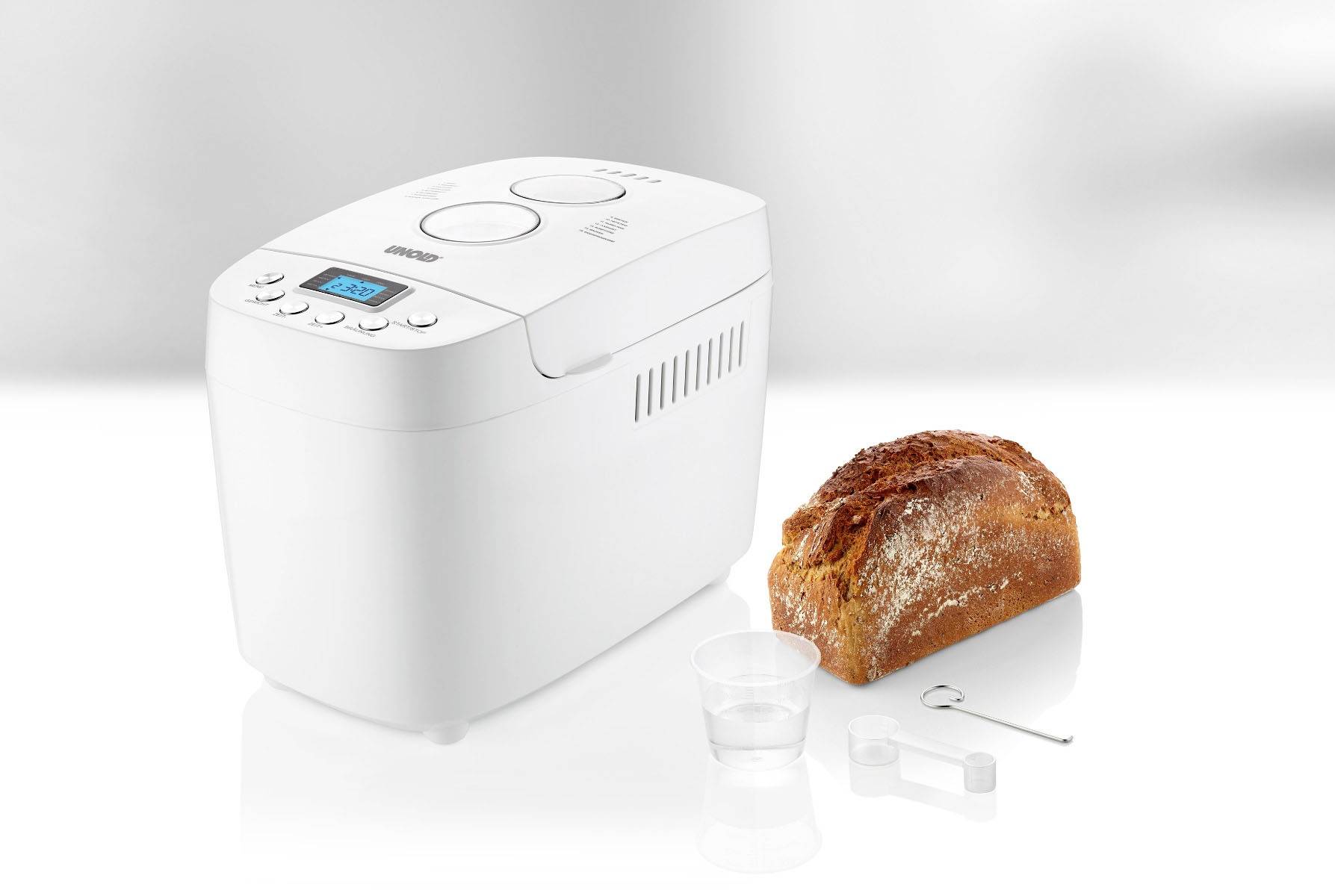 Ein weißer Brotbackautomat neben einem frisch gebackenen Laib Brot, einem Messbecher und einem Haken. Das Display zeigt die Betriebsanzeige.