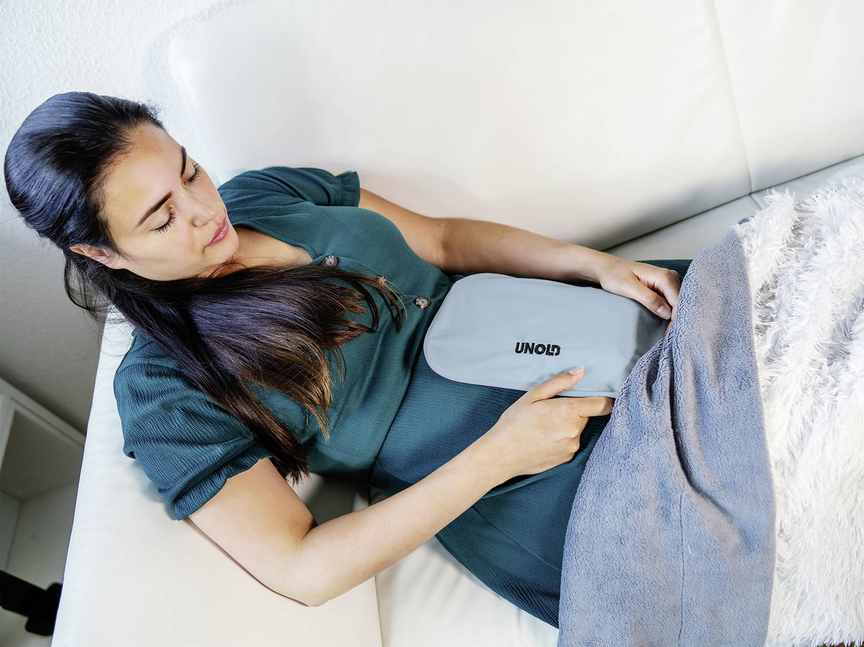 Frau in grüner Kleidung entspannt sich auf einem Sofa, hält ein Wärme- oder Massagegerät unter einem grauen Tuch auf ihrem Bauch.