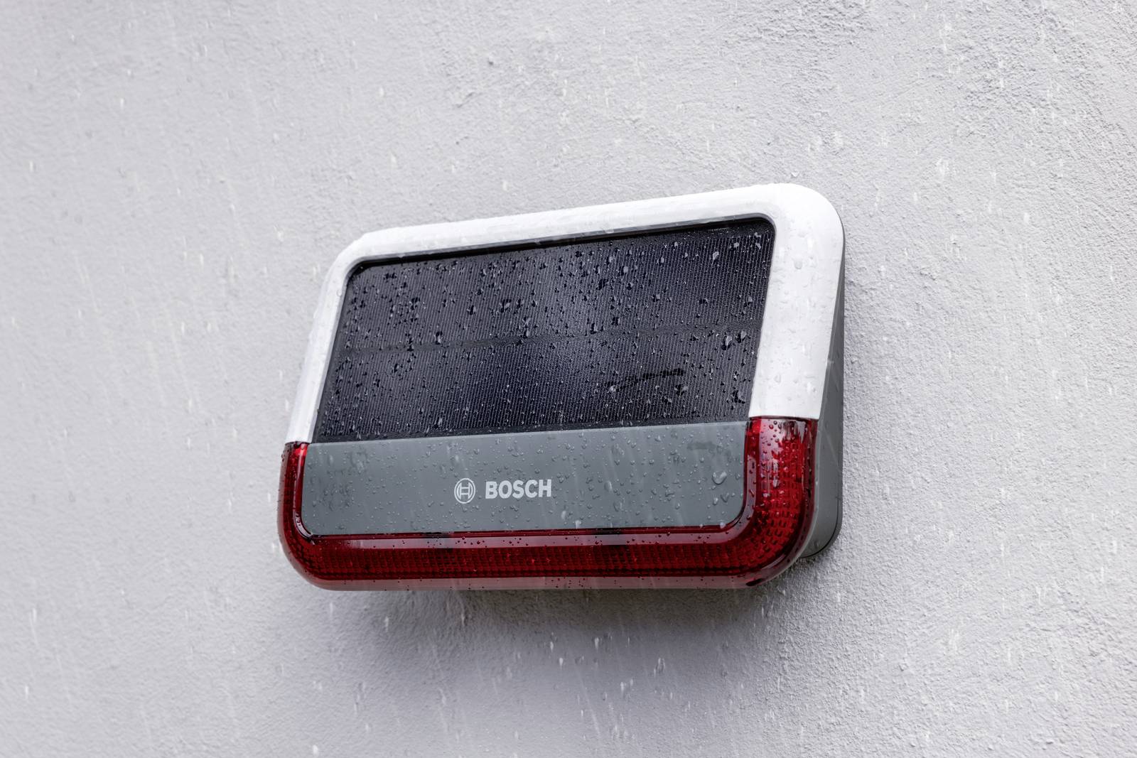 Kompakte, rechteckige Außenalarmanlage an einer Wand, mit 'Bosch' Logo. Sie zeigt Regentropfen, was Wetterfestigkeit andeutet.