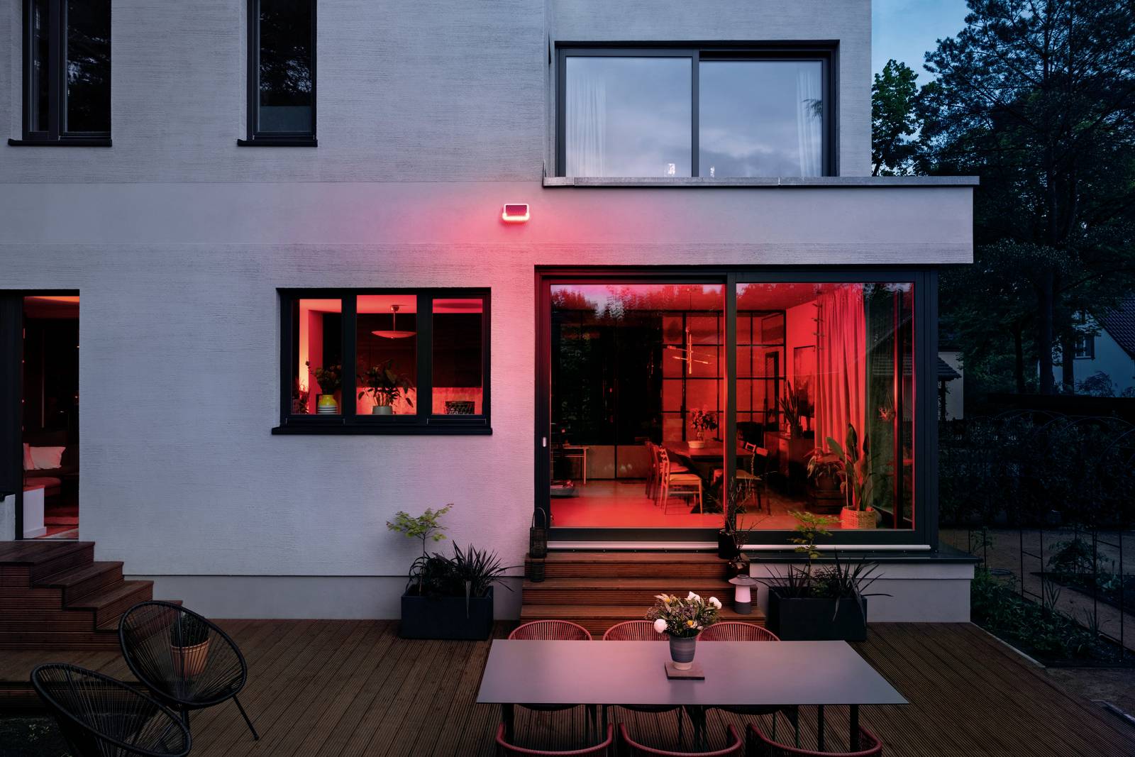 Ein modernes Haus bei Nacht mit einem großen Fenster, durch das rotes Licht scheint. Draußen stehen ein Tisch und Stühle auf einer Terrasse.