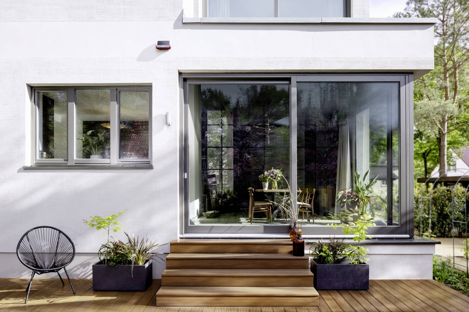 Modernes Haus mit großen Glas-Schiebetüren, die zu einer Terrasse mit Stufen führen. Pflanzen in Töpfen dekorieren den Bereich.