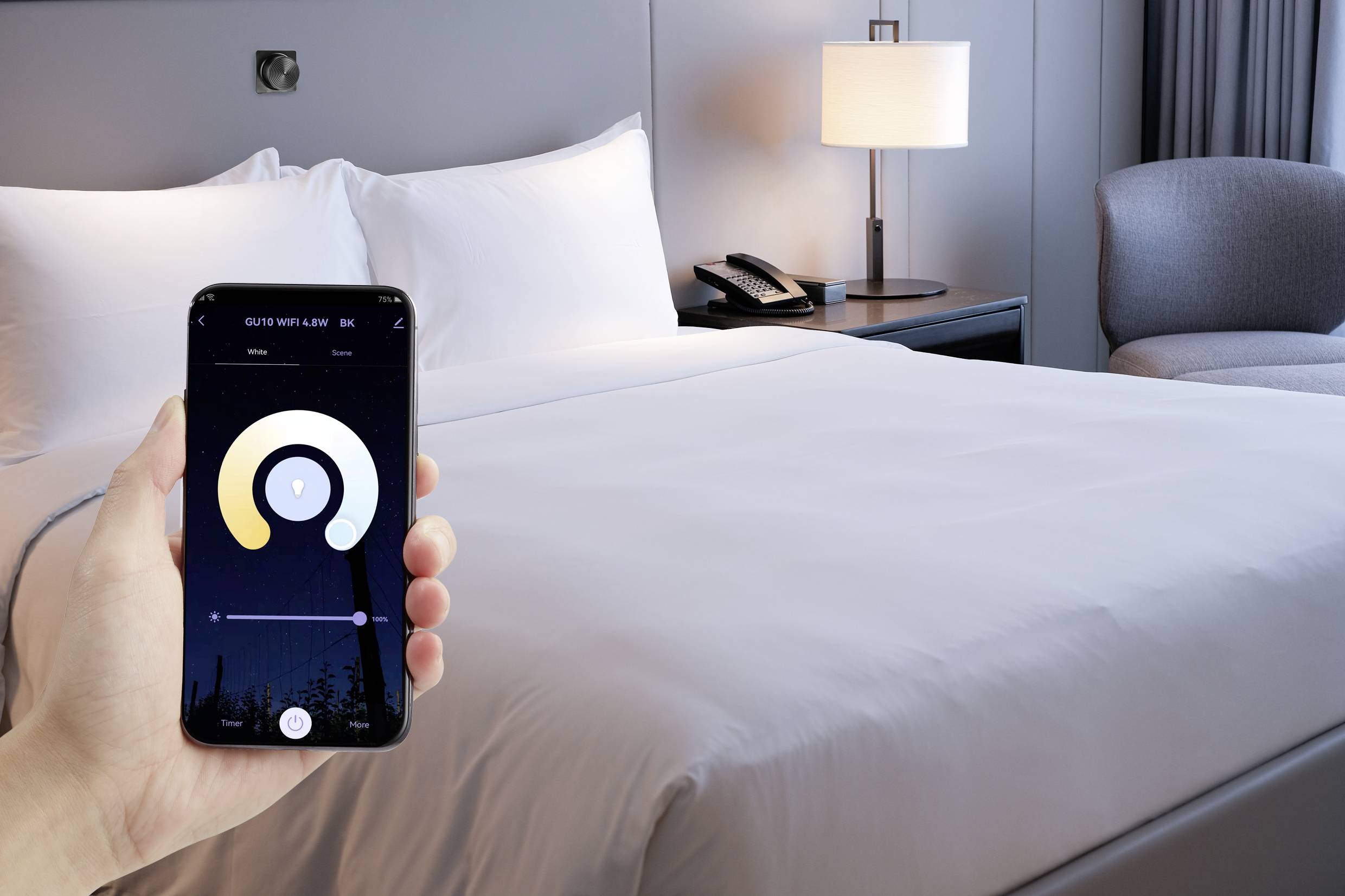 Eine Hand hält ein Smartphone, das eine Smart-Home-App zeigt, vor einem Hotelbett. Im Hintergrund Nachttisch mit Lampe und Telefon.