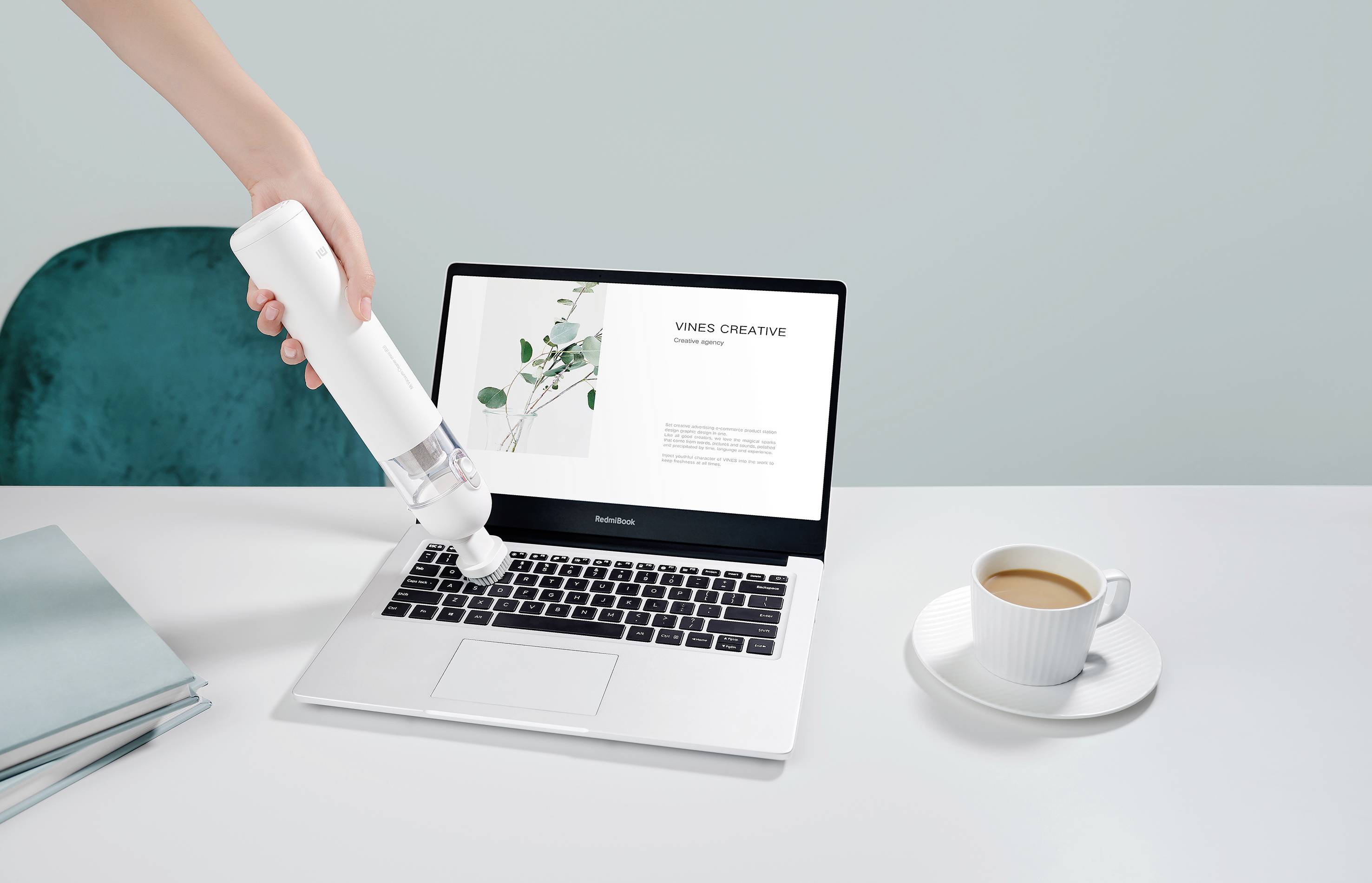 Eine Hand verwendet einen weißen Staubsauger, um einen Laptop zu reinigen. Daneben ein Kaffeetasse und Bücher auf einem Tisch.