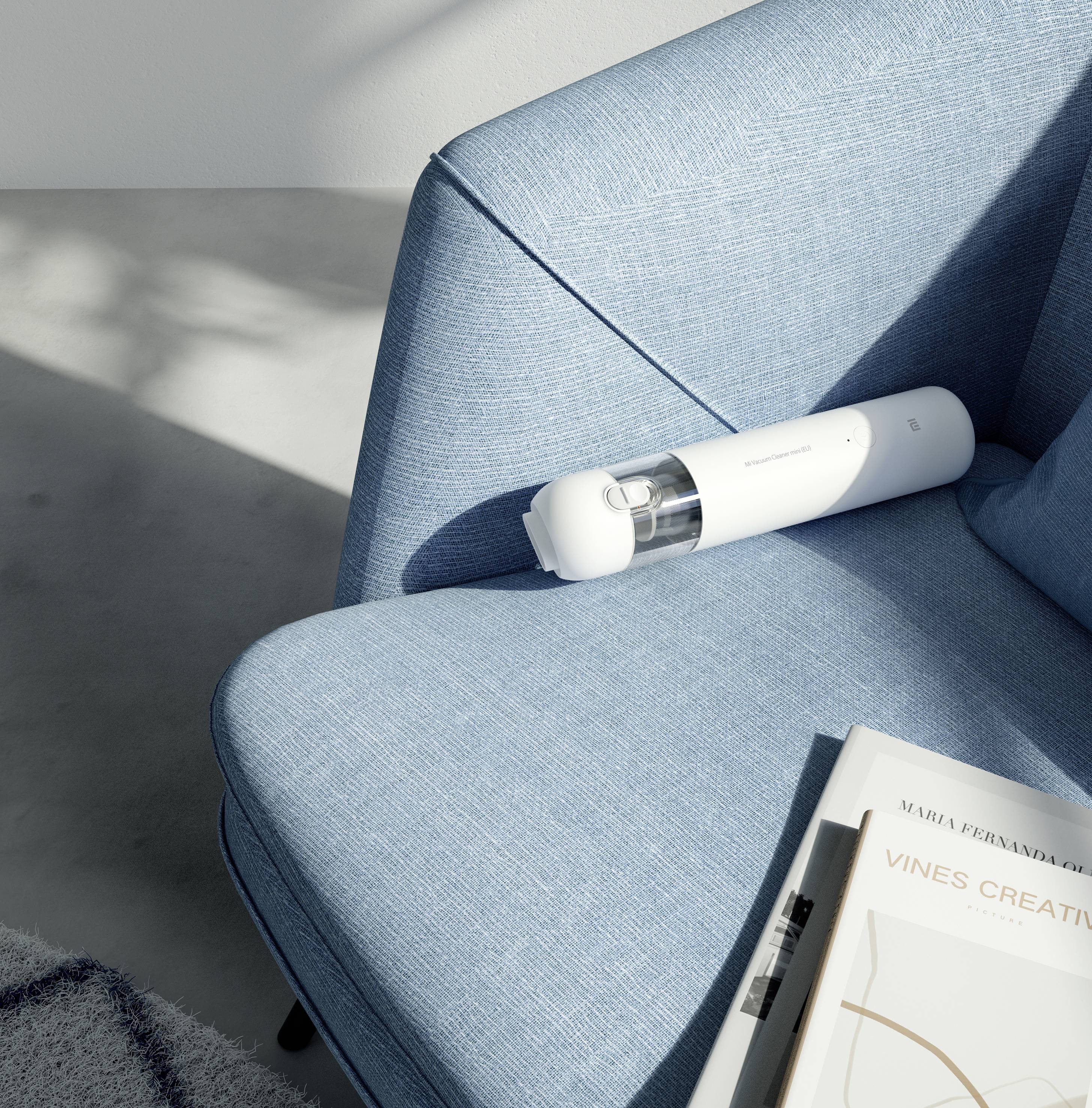 Ein kleiner, weißer Handstaubsauger liegt auf einem blauen Sofa. Neben dem Staubsauger liegt ein Buch mit weißem Einband.