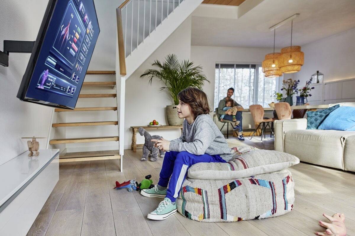 Ein Kind spielt Videospiele auf einem Fernseher in einem gemütlichen Wohnzimmer; Eltern sitzen im Hintergrund auf der Couch.