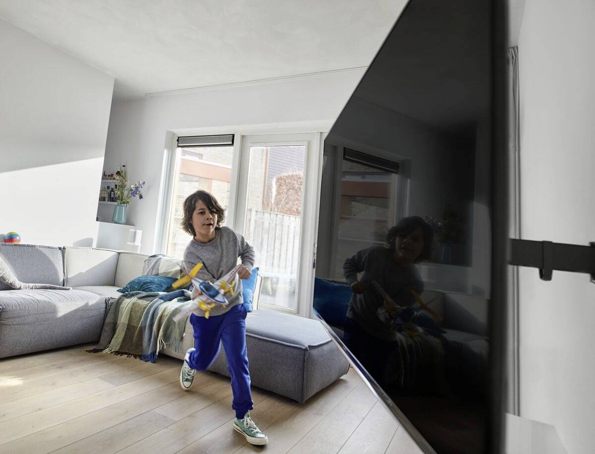 Ein Kind rennt durch ein Wohnzimmer mit einem Spielzeug in der Hand. Im Hintergrund sind ein Sofa, ein Tisch und große Fenster zu sehen.