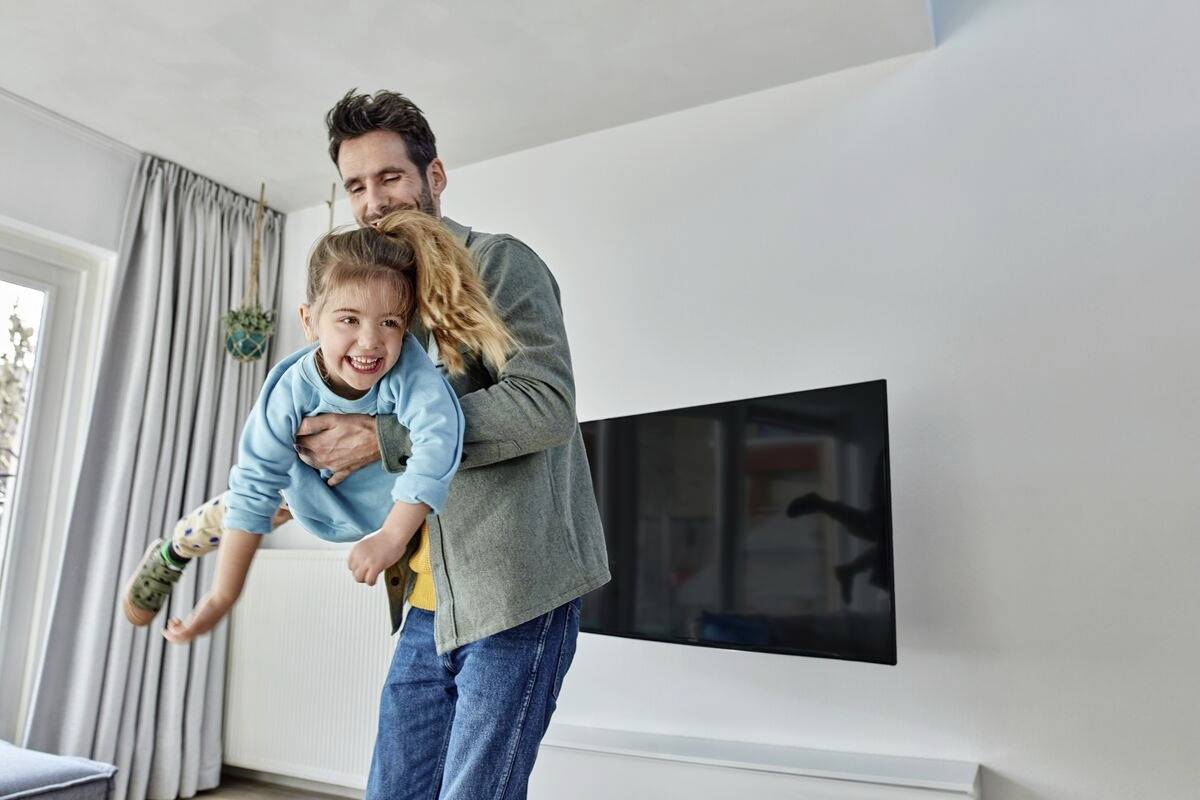 Ein Mann hebt ein lachendes Kind in einem Wohnzimmer hoch. Im Hintergrund hängt ein Fernseher an der Wand.