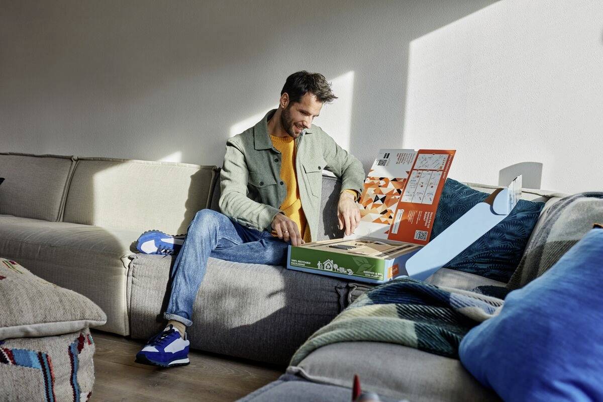 Ein Mann sitzt auf einem Sofa und öffnet eine Pizzaschachtel. Er wirkt entspannt und lächelt, während Sonnenlicht den Raum erhellt.