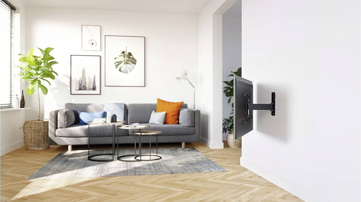 Moderner, heller Wohnraum mit grauem Sofa, bunten Kissen und Pflanzen. An der Wand ein montierter Bildschirm. Holzboden und große Fenster.