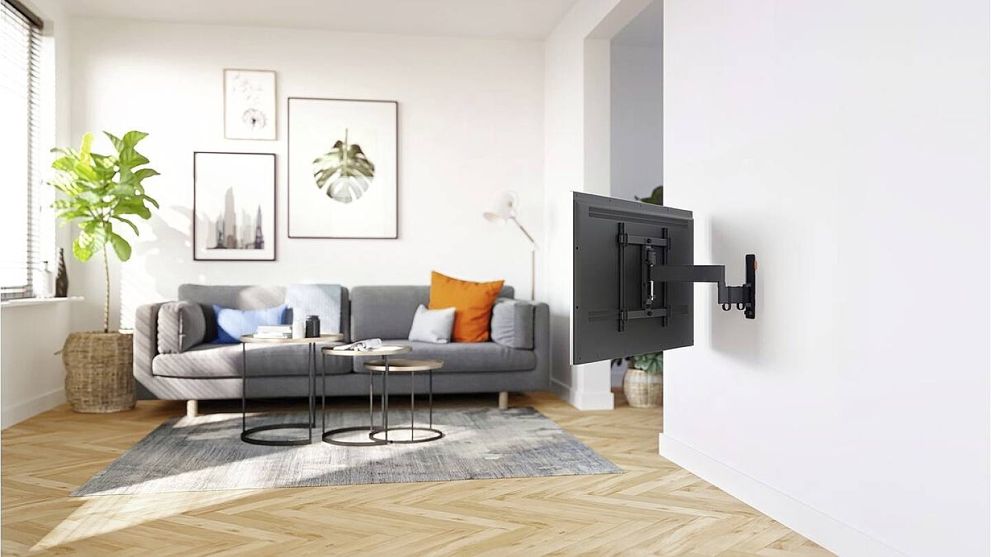 Helles Wohnzimmer mit grauem Sofa, bunten Kissen, Pflanzen, und einem an der Wand montierten Fernseher auf einem schwenkbaren Arm.