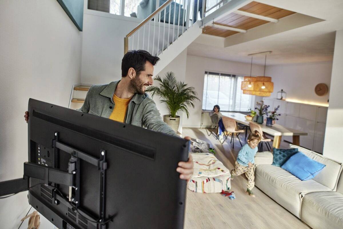 Ein Mann hebt einen großen Fernseher hoch, während ein Kind im Wohnzimmer spielt. Ein anderer erwachsener Mensch sitzt auf dem Sofa.