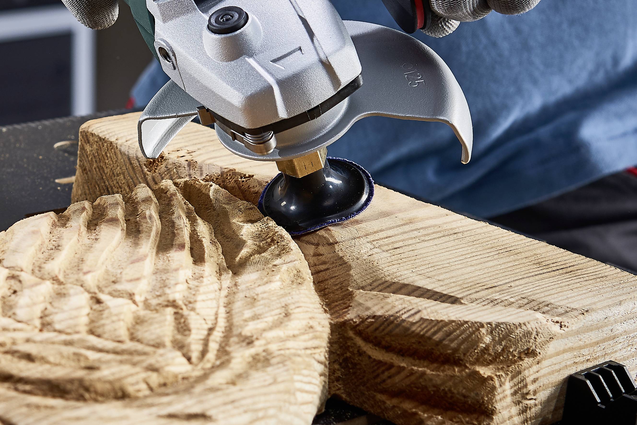Ein Holzstück wird mit einem Winkelschleifer bearbeitet. Die Oberfläche zeigt bearbeitete Muster, was auf eine Holzschnitzarbeit hindeutet.